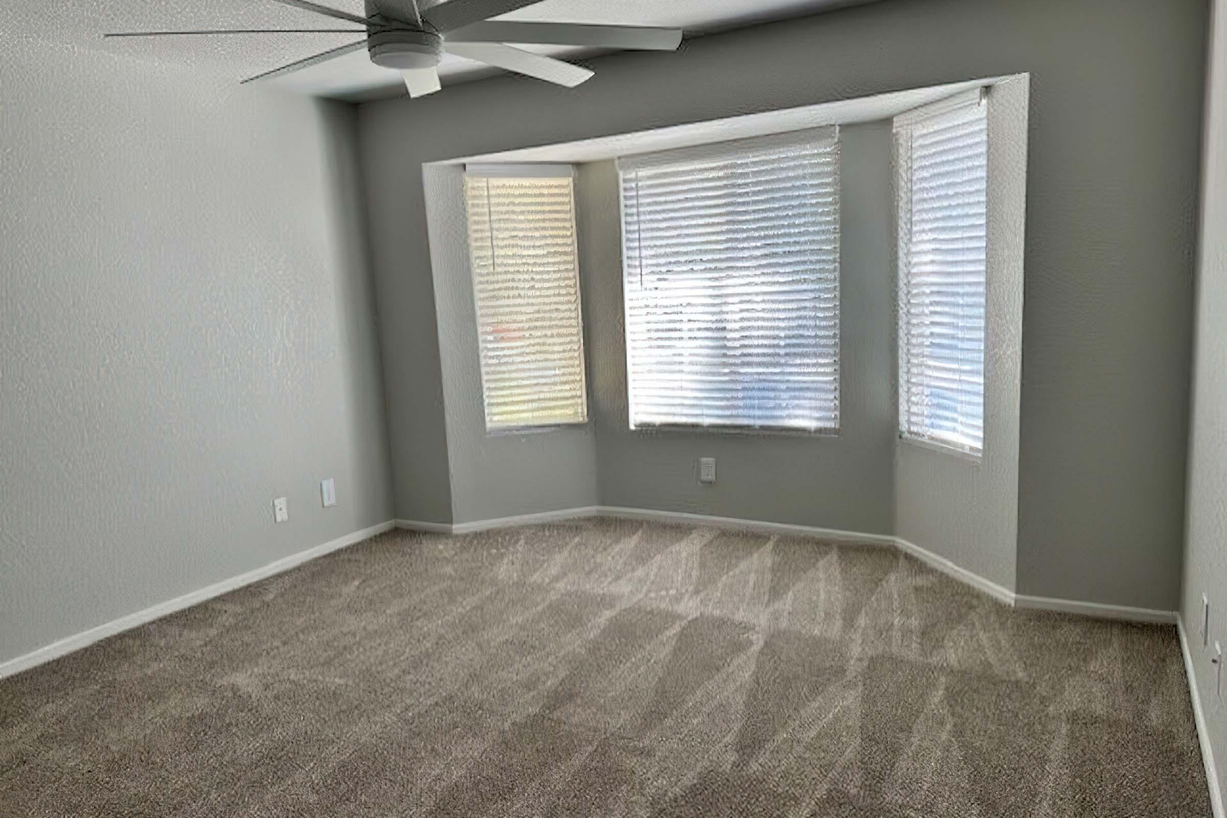 A bright, empty room with light gray walls and a plush beige carpet. The room features large bay windows with sheer blinds allowing natural light to filter in. A ceiling fan is mounted above, adding a modern touch to the simple, spacious layout.