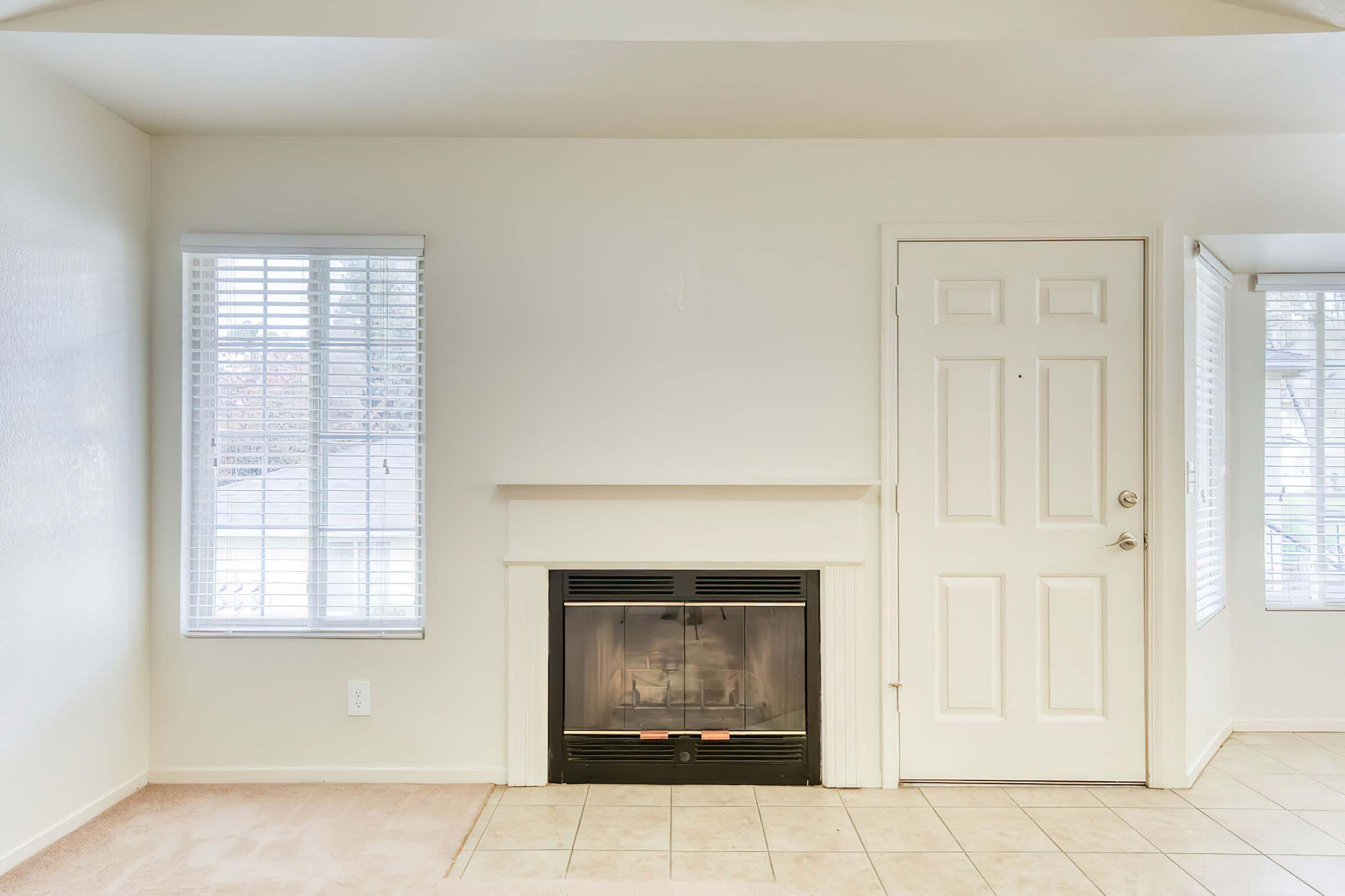 A bright, minimally furnished room featuring a white wall with two windows covered by blinds. There's a simple fireplace with a glass front in the center, and a white door on the right side. The flooring is a combination of tiles and carpet, creating a clean and open atmosphere.