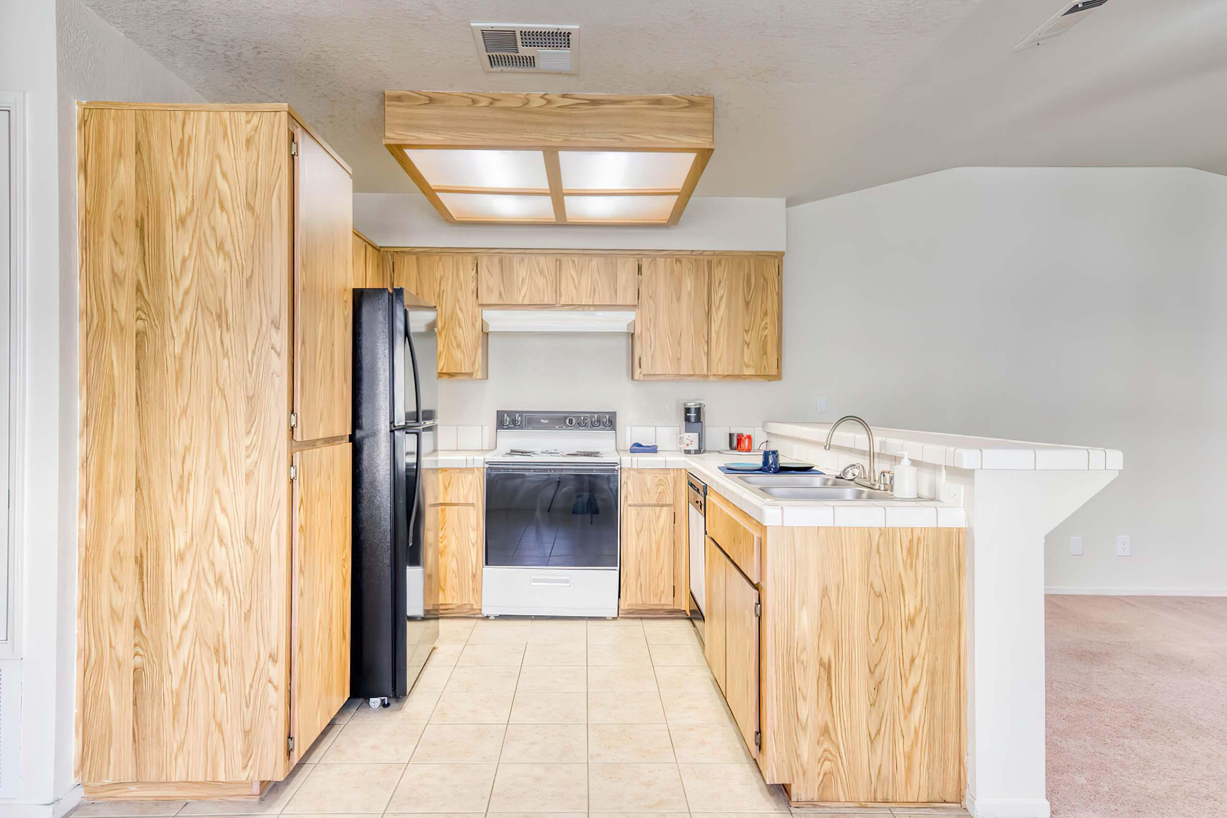 Bright kitchen with wooden cabinets and light-colored countertops. Features a black refrigerator, a white stove, and a sink. The floor is tiled, and there's a small bar area visible. Natural light comes from an overhead fixture. The walls are painted in a light color, creating an airy atmosphere.