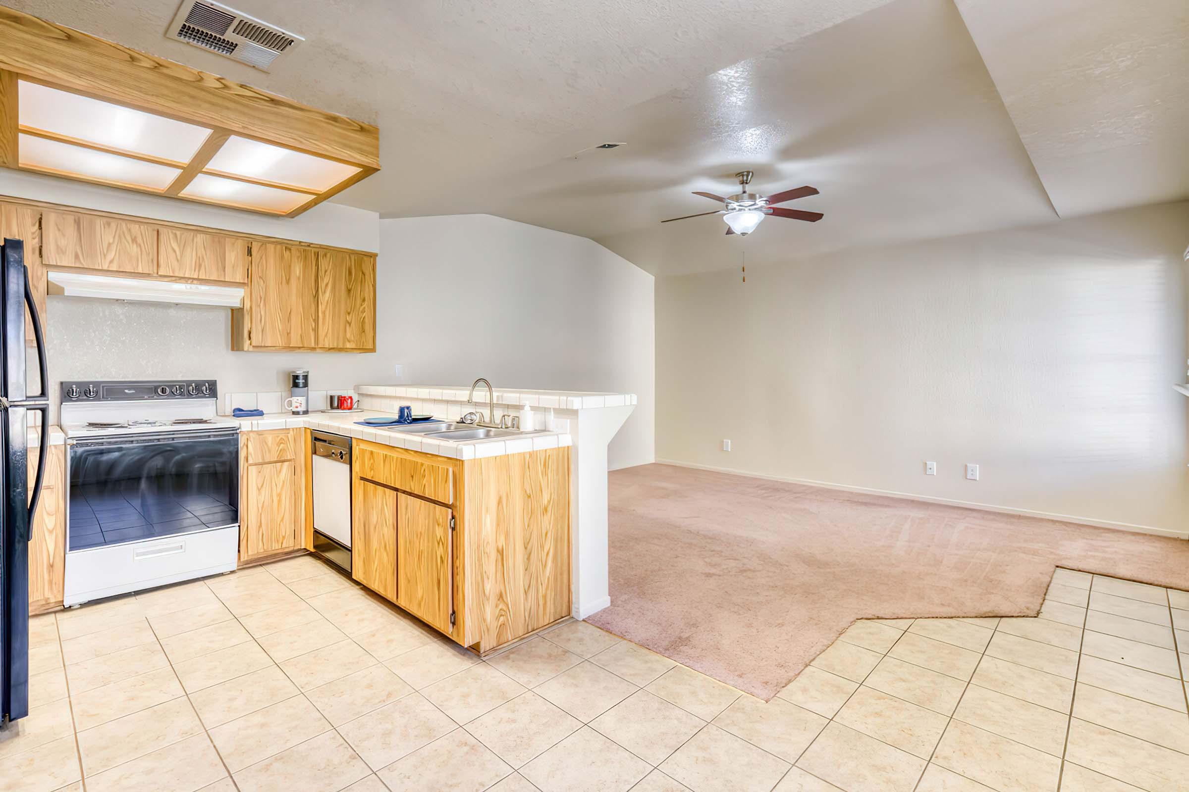 A spacious kitchen with wooden cabinets, a stove, and sink, leading into a living area with light-colored carpet and a ceiling fan. The kitchen has tile flooring and ample natural light from a nearby window. The living area features neutral walls, creating a bright and inviting space.