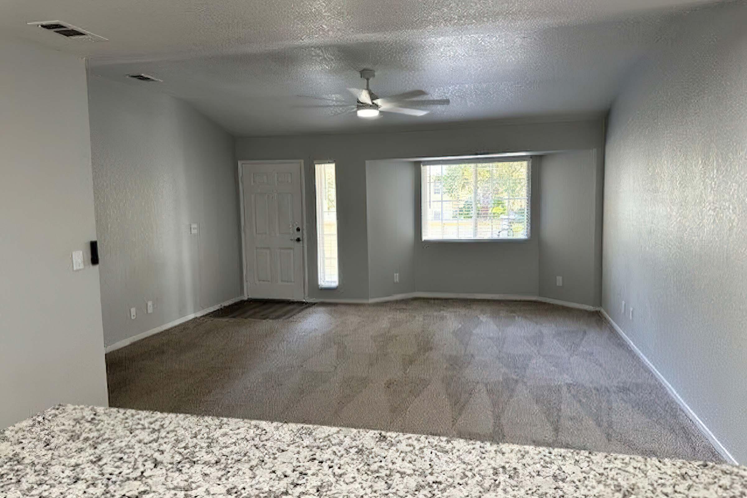 A spacious living room with light gray walls and carpet. It features a ceiling fan and a large window allowing natural light. A front door is visible on the left, and a small entry area is present. The view includes a part of a kitchen countertop in the foreground.
