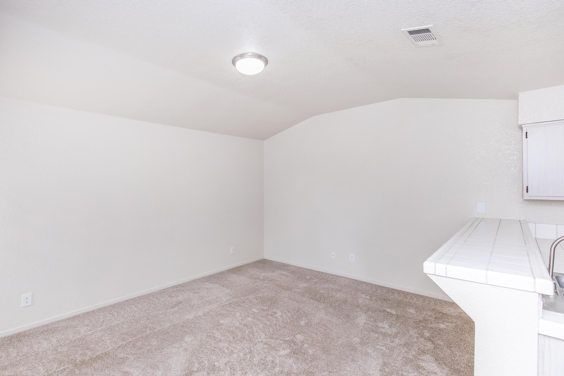 A spacious, empty room with beige carpet and light-colored walls. The ceiling is slightly arched, and there is a small ceiling light fixture. In the background, a kitchenette with white cabinets and a tile countertop is partially visible. The space appears bright and well-lit.