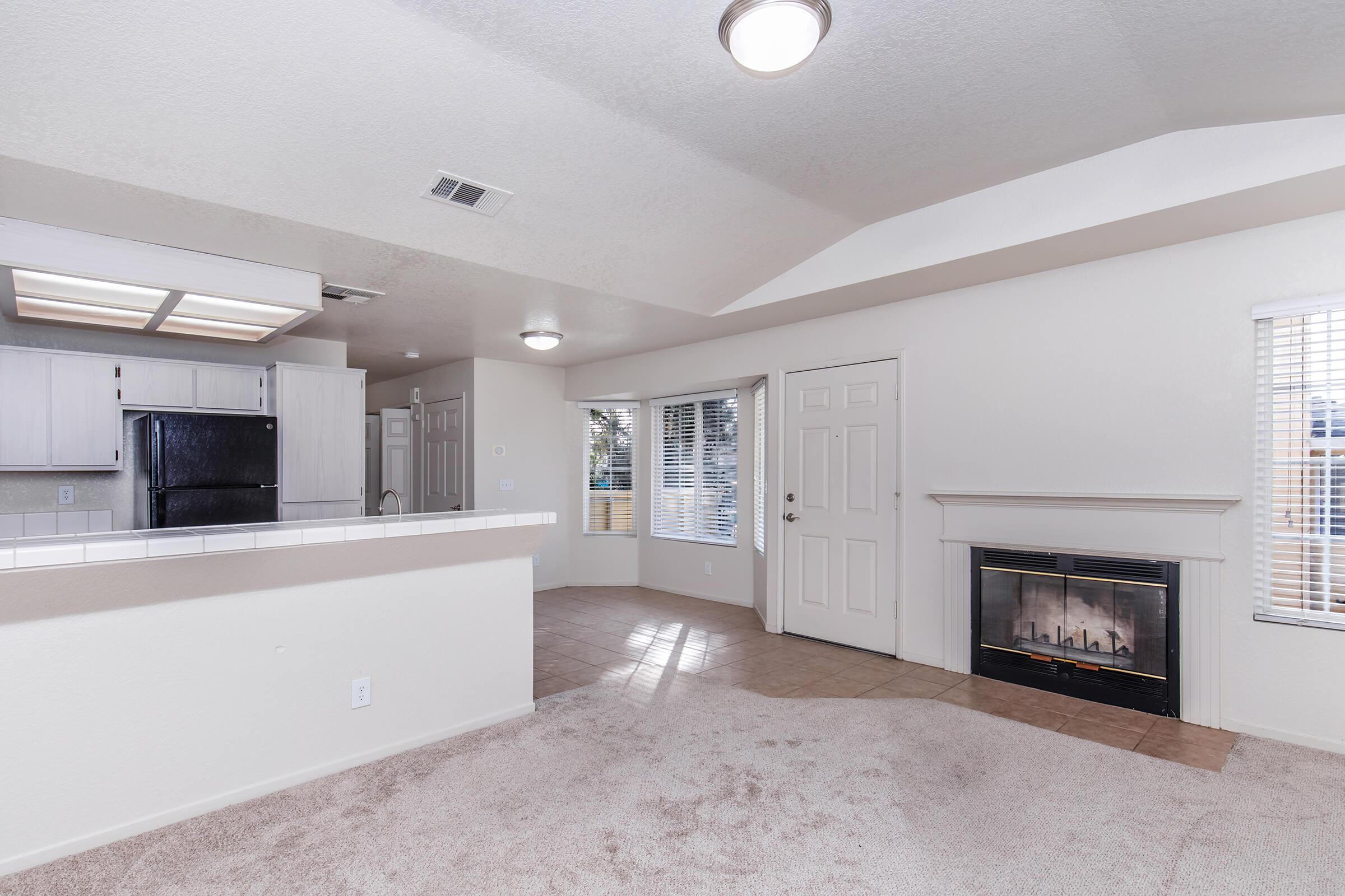 Bright, open living space featuring a cozy fireplace, light-colored walls, and large windows with blinds. The kitchen area is visible with a bar countertop, and there's a carpeted floor enhancing the warmth of the room. The layout provides a comfortable and inviting atmosphere.