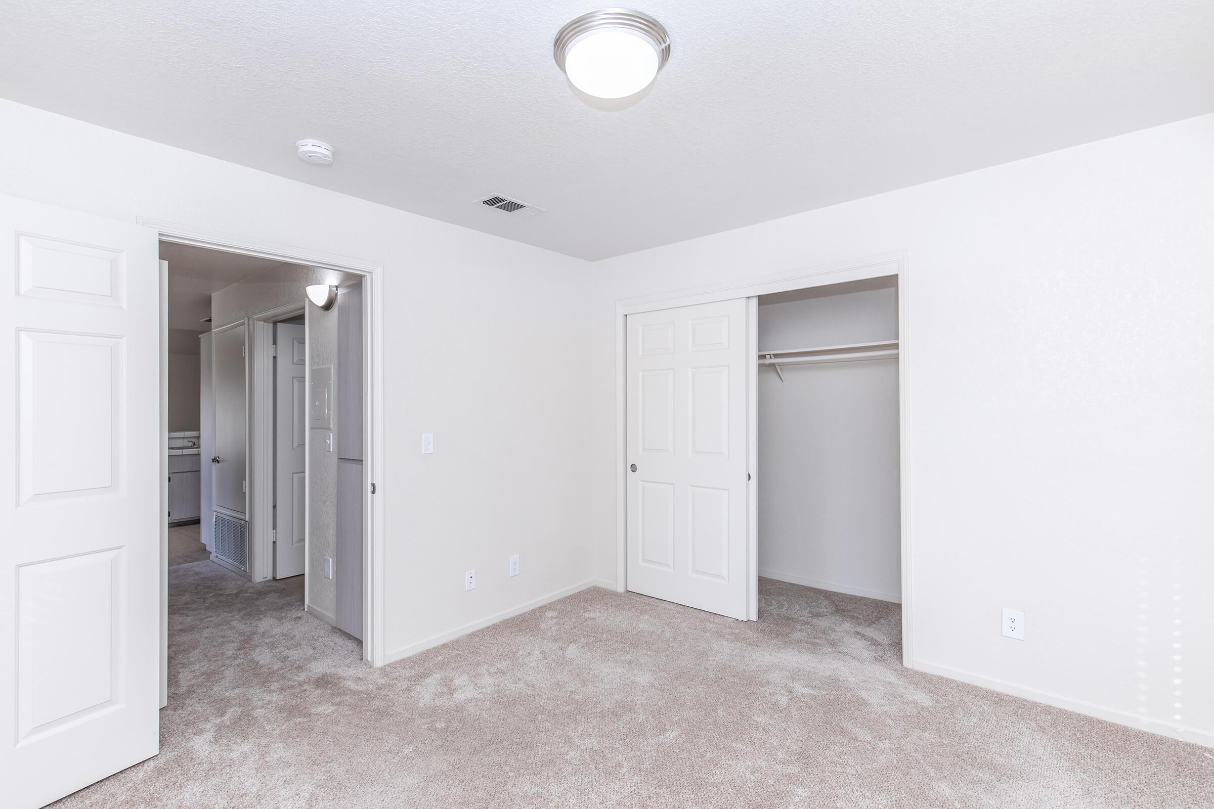 A well-lit, empty room with beige carpeting and white walls. There are two doors leading to adjacent spaces, one of which is a closet. The ceiling features a circular light fixture, enhancing the spacious feel of the area. The overall design is simple and modern.