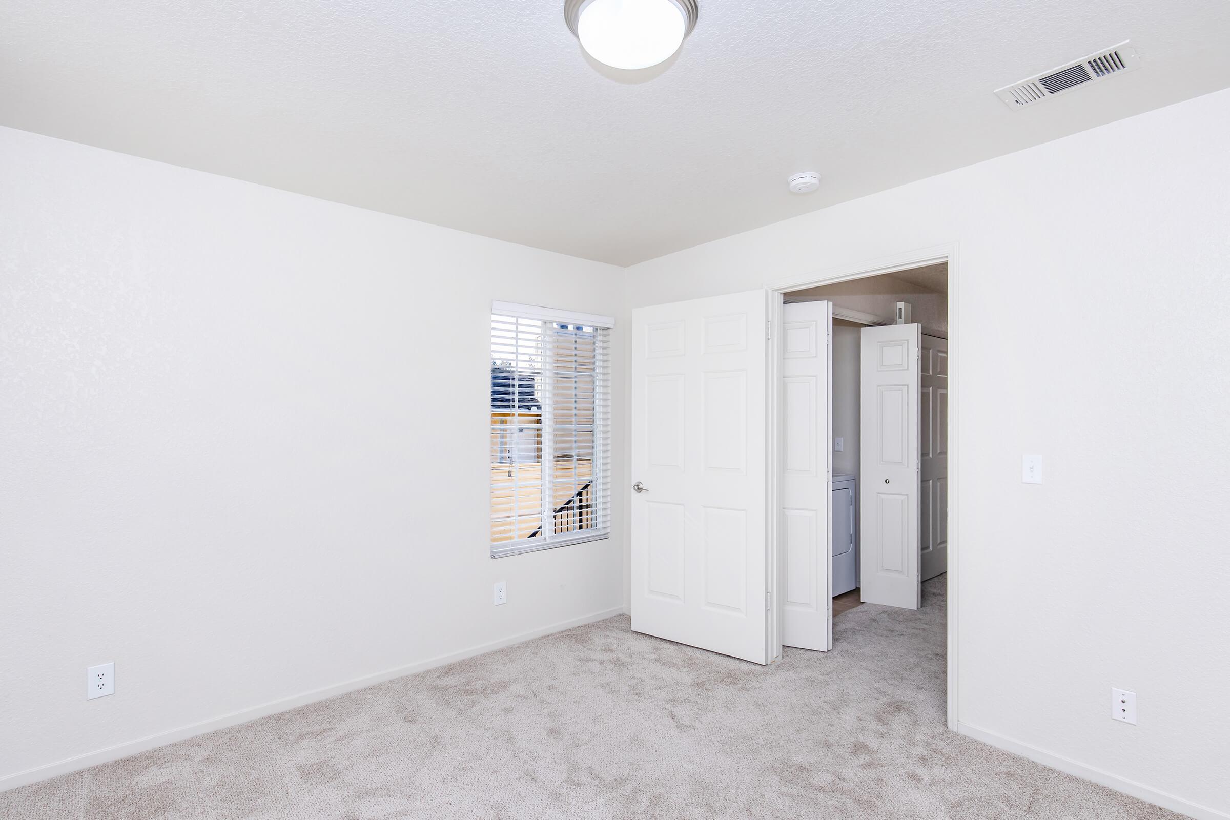 A well-lit, empty room with beige carpet, white walls, and a ceiling light. There are two closed doors, one leading out of the room and another possibly to a closet. A window with horizontal blinds allows natural light in, and there is a smoke detector on the ceiling.