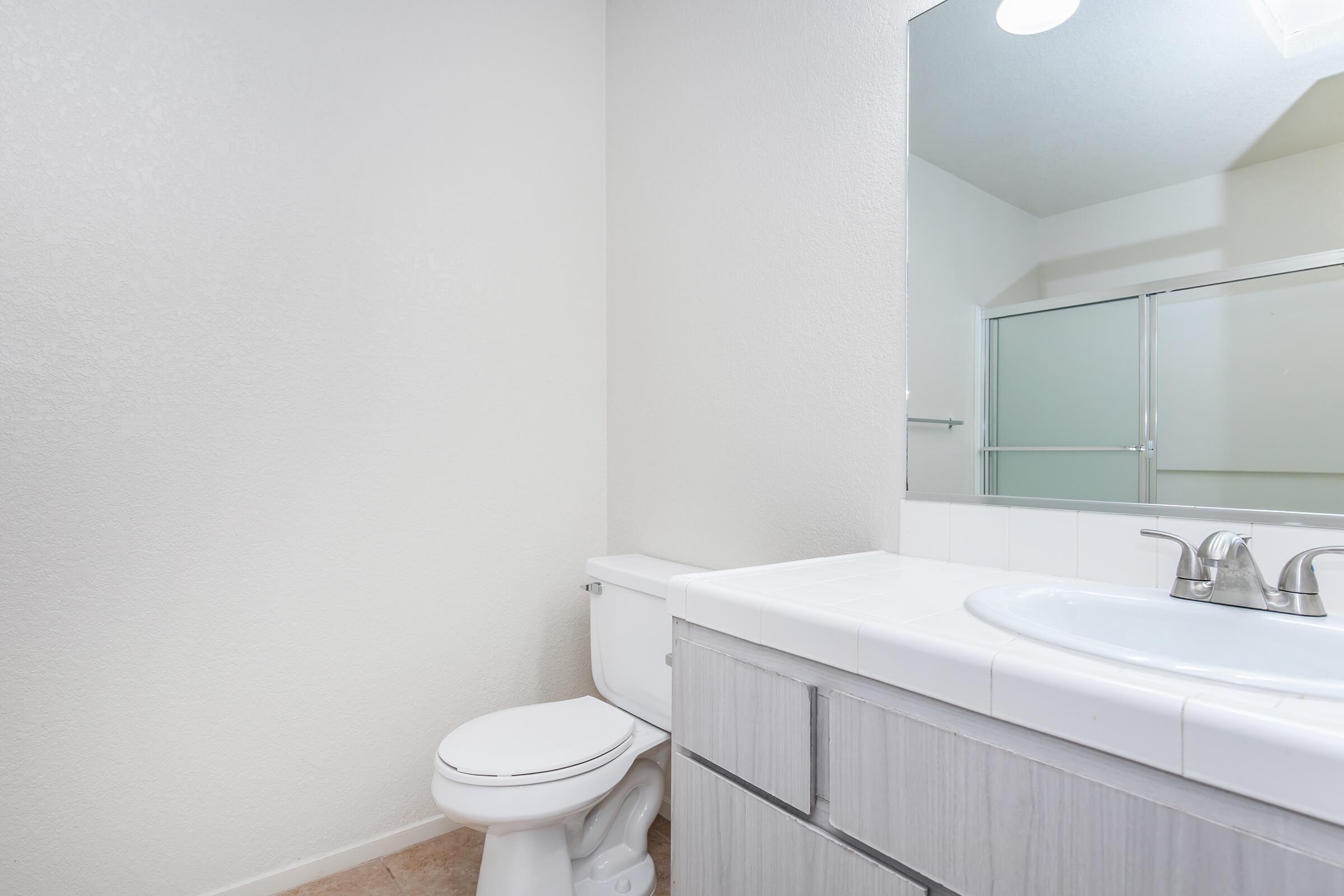 A clean bathroom featuring a white toilet, a simple sink with a silver faucet, and a large mirror. The walls are light-colored, and a glass shower enclosure is visible in the background, creating a bright and spacious atmosphere. The flooring is tile, contributing to a modern look.