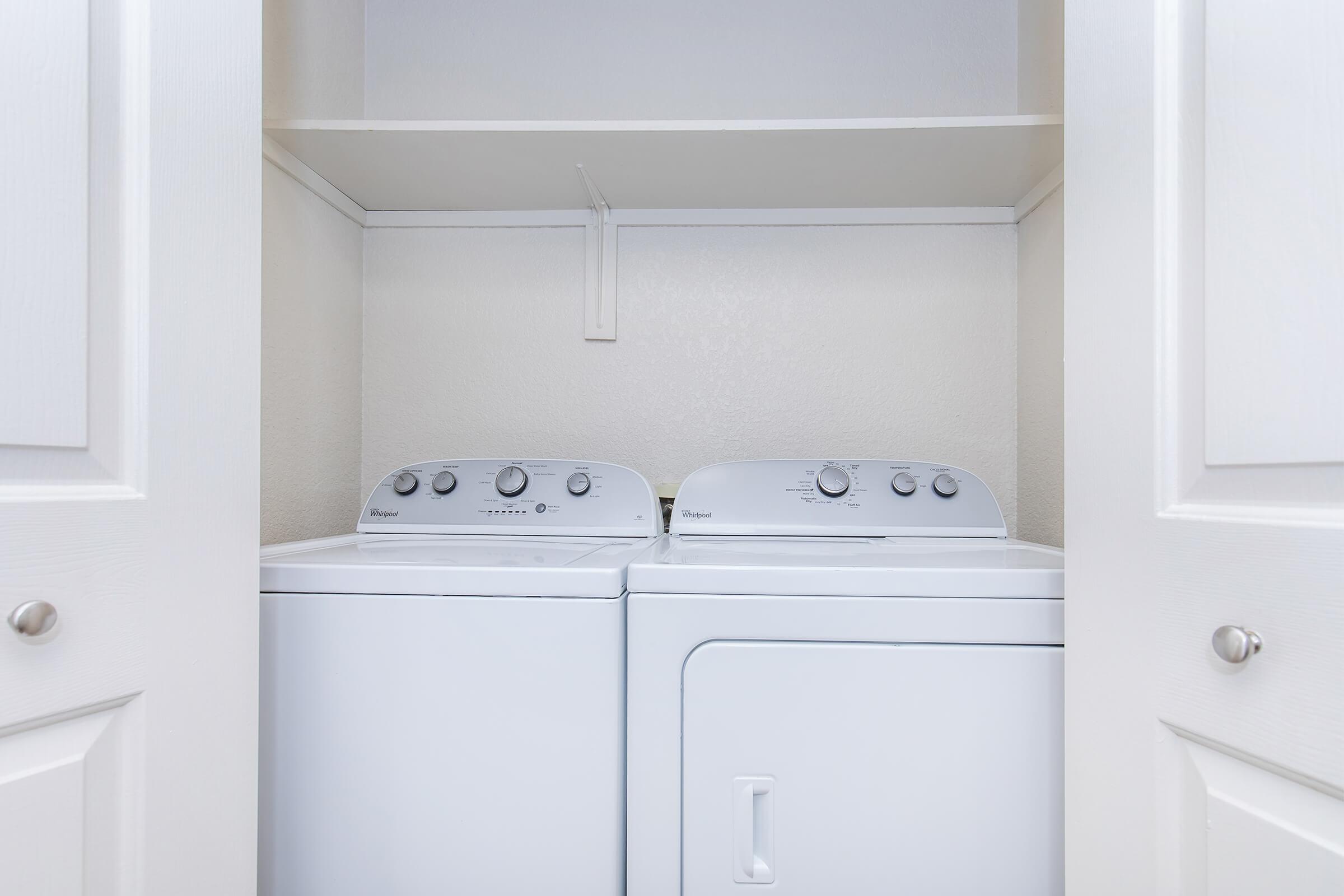 A laundry area featuring a white washing machine and dryer side by side, with controls visible on the top. The appliances are housed in a small closet space with white doors and a shelf above. The walls are a light color, creating a clean and organized appearance.