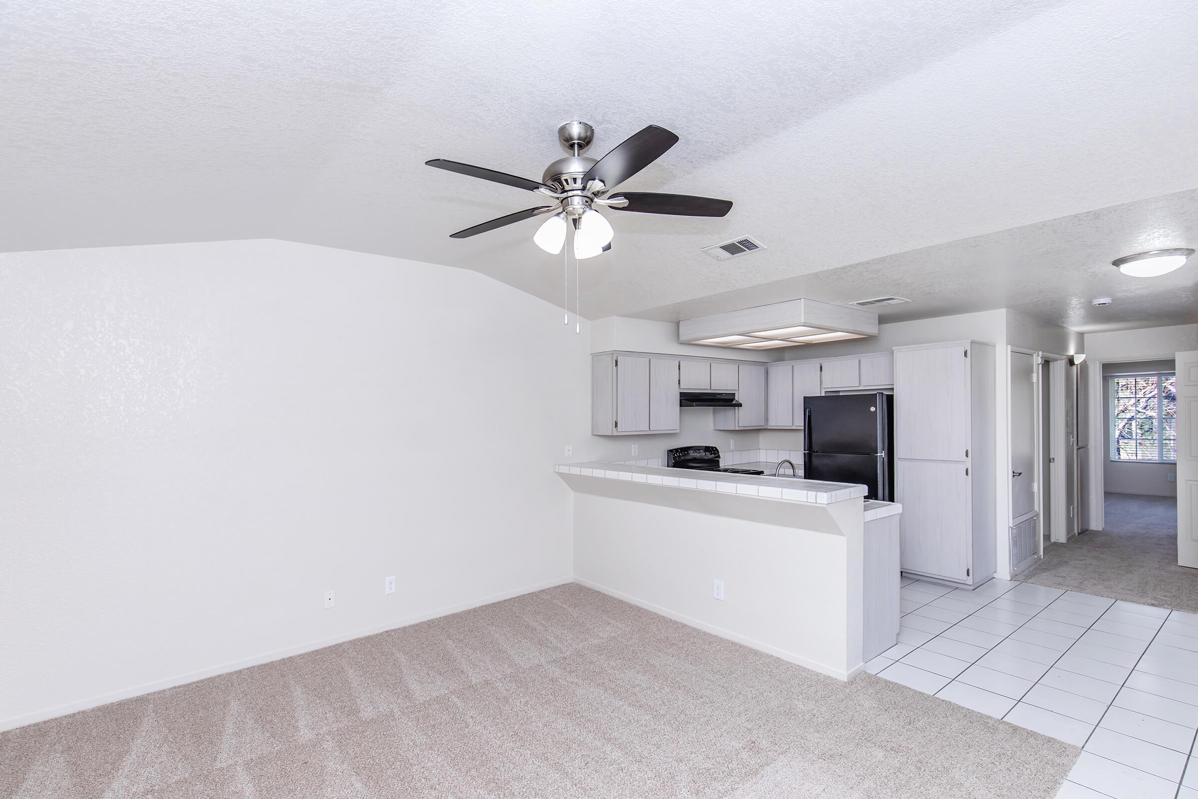 Open-concept living area featuring a ceiling fan, carpeted floor, and a kitchen area with modern appliances. The space includes light-colored walls and tiles, providing a bright and airy atmosphere, with an adjoining hallway leading to additional rooms.