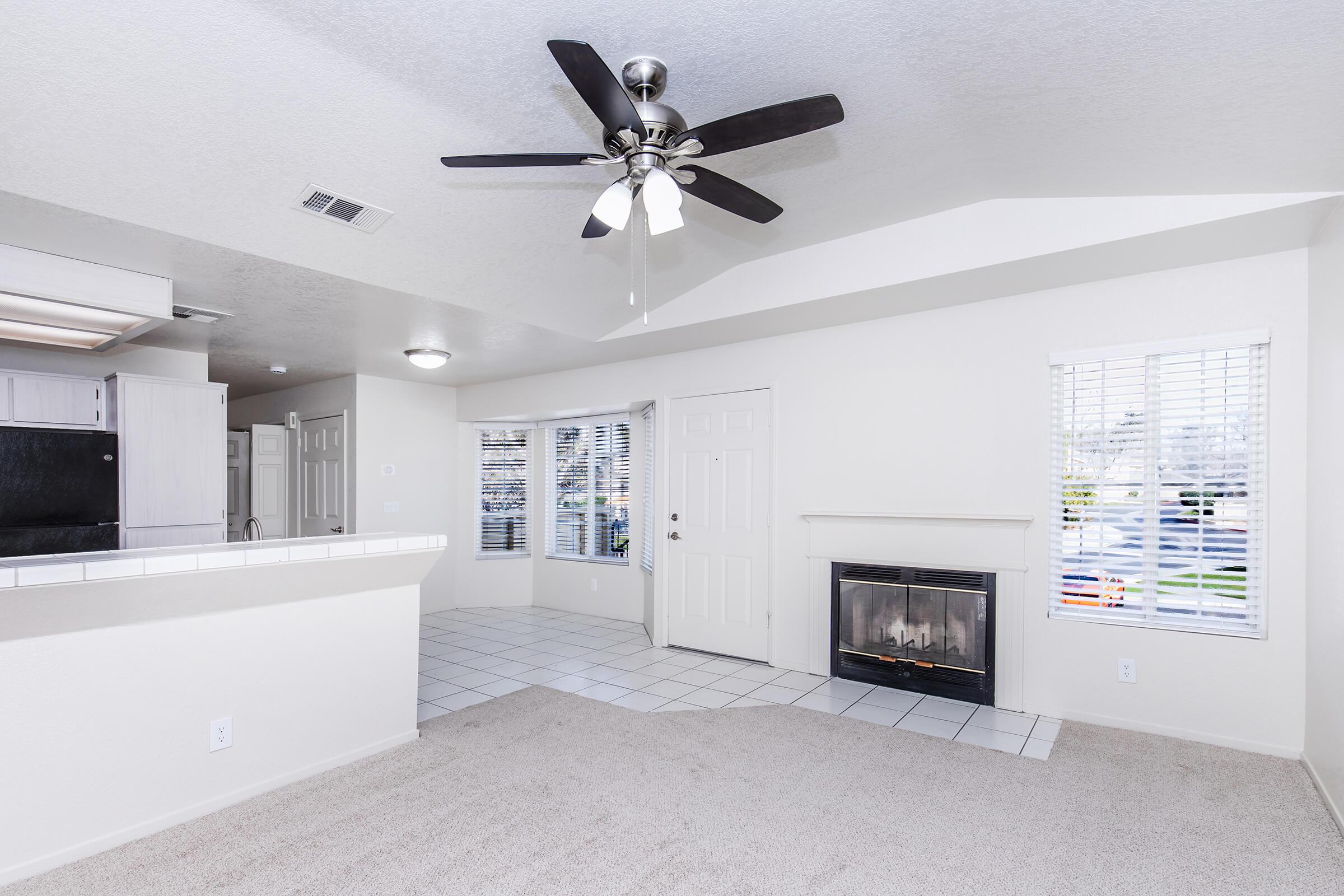 Bright and spacious living area featuring a ceiling fan, carpet flooring, and tiled section. Includes a corner fireplace and large windows with blinds, providing natural light. An open layout connects to a kitchen area with white cabinetry.