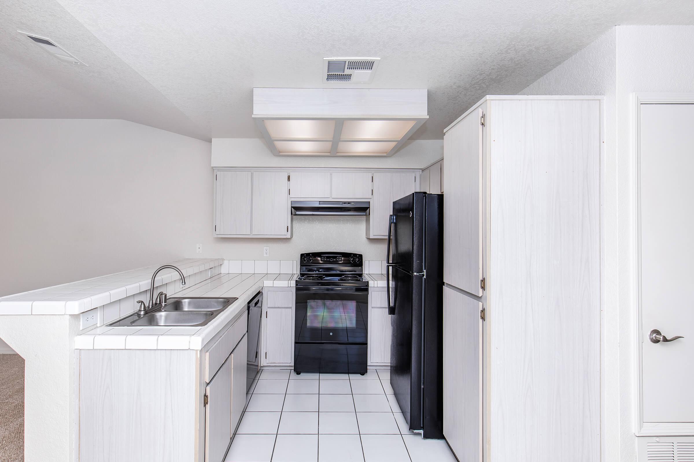 A modern kitchen featuring white cabinetry, a black stove and refrigerator, and a double sink. The floor is tiled in white, and there's a coffered ceiling with lights above. The kitchen has an open layout leading to an adjacent room.