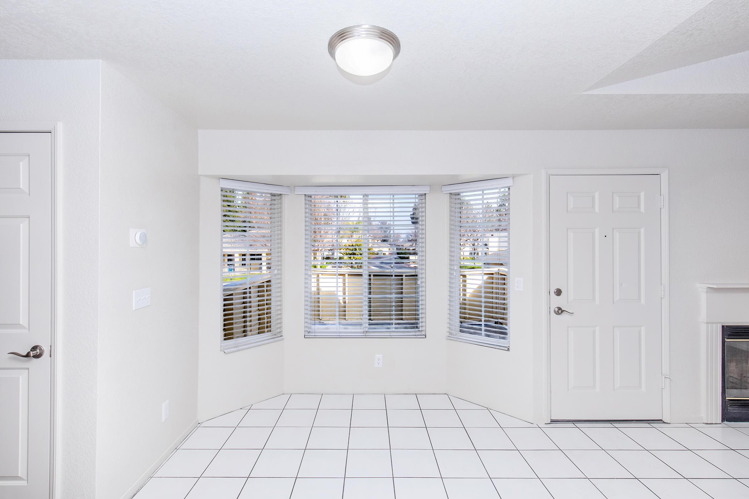A bright, empty interior room featuring white walls and tile flooring. It has large windows with blinds on two sides, letting in natural light. A white door is visible on one side, with a small area for a fireplace. The overall space is clean and minimalistic.