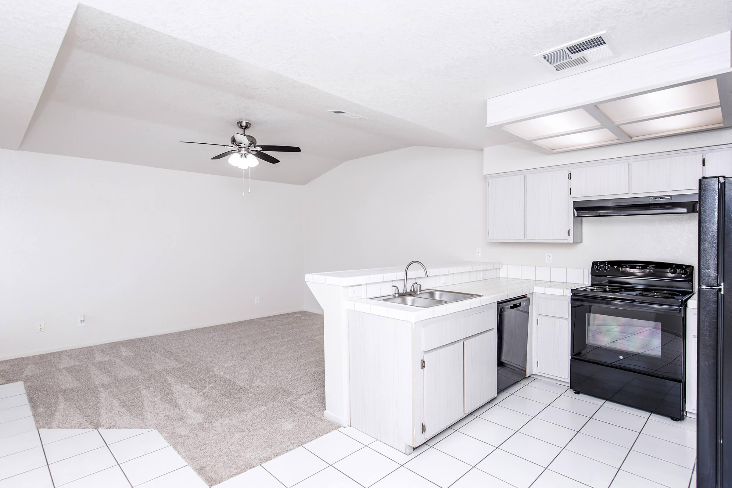 A spacious, empty kitchen and living area with light-colored walls. The kitchen features a ceiling fan, white cabinetry, a double sink, a black stove, and a refrigerator. The flooring is a mix of tile and carpet. The room has a bright, airy feel with ample natural light.