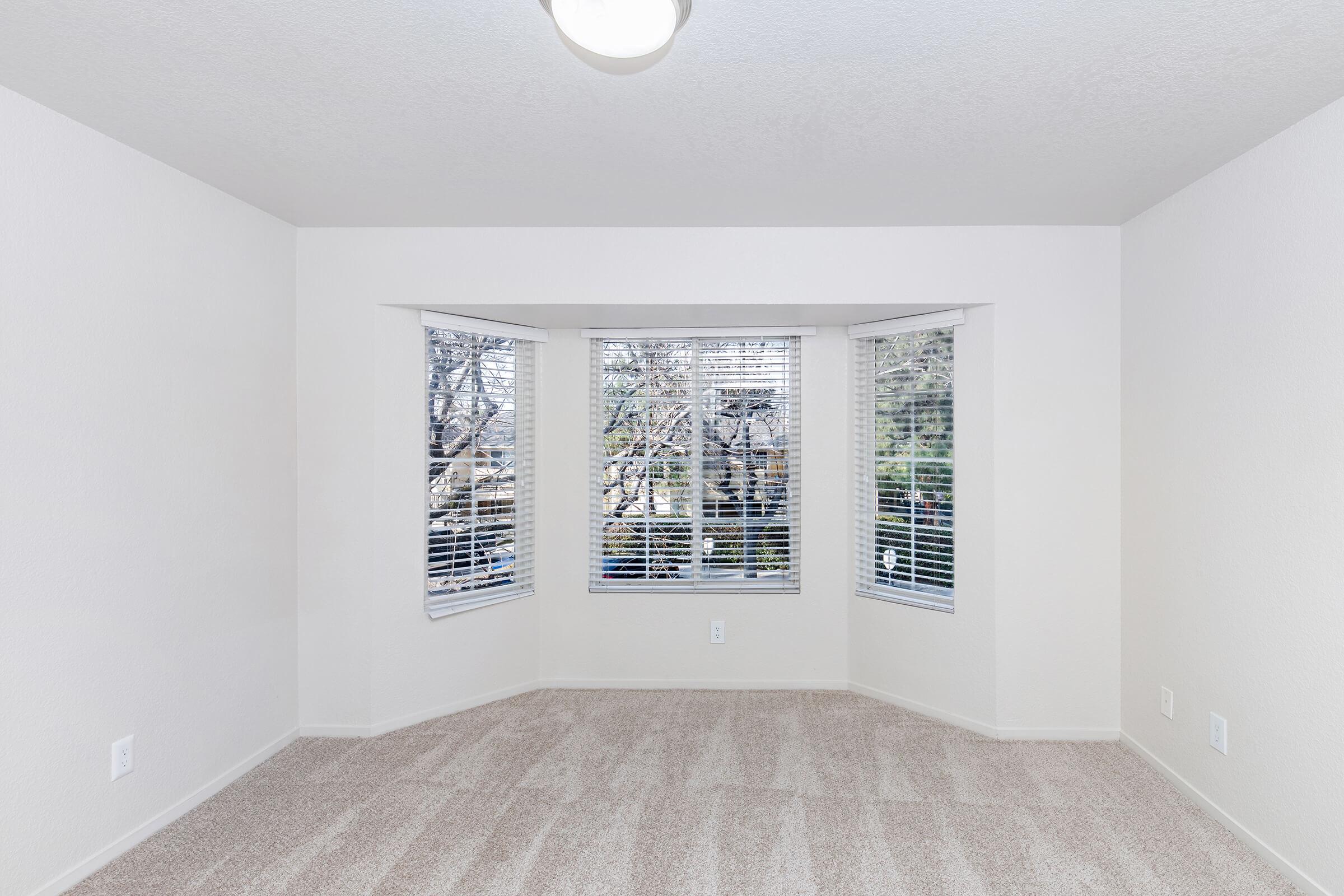 A bright, empty room features light beige carpet and white walls. Three large windows with closed blinds allow natural light to illuminate the space. The room has a simple ceiling light fixture and is devoid of furniture, providing a clean, minimalist aesthetic.