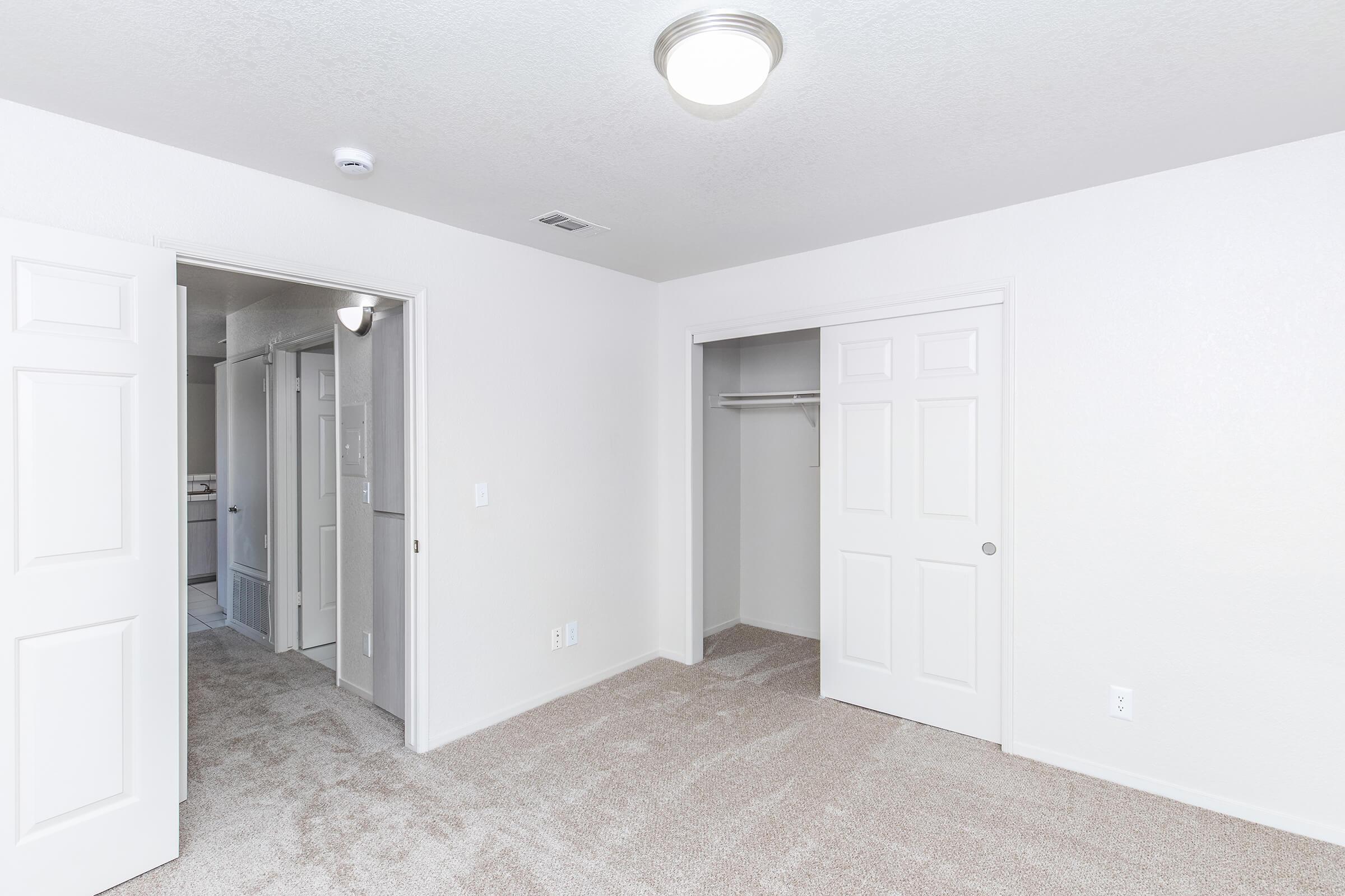 Bright, empty room with light-colored walls and carpet. Features two white doors: one leading to a closet and another to a hallway. The space is well-lit and clean, creating an open and airy atmosphere. Ideal for personalizing or decorating.