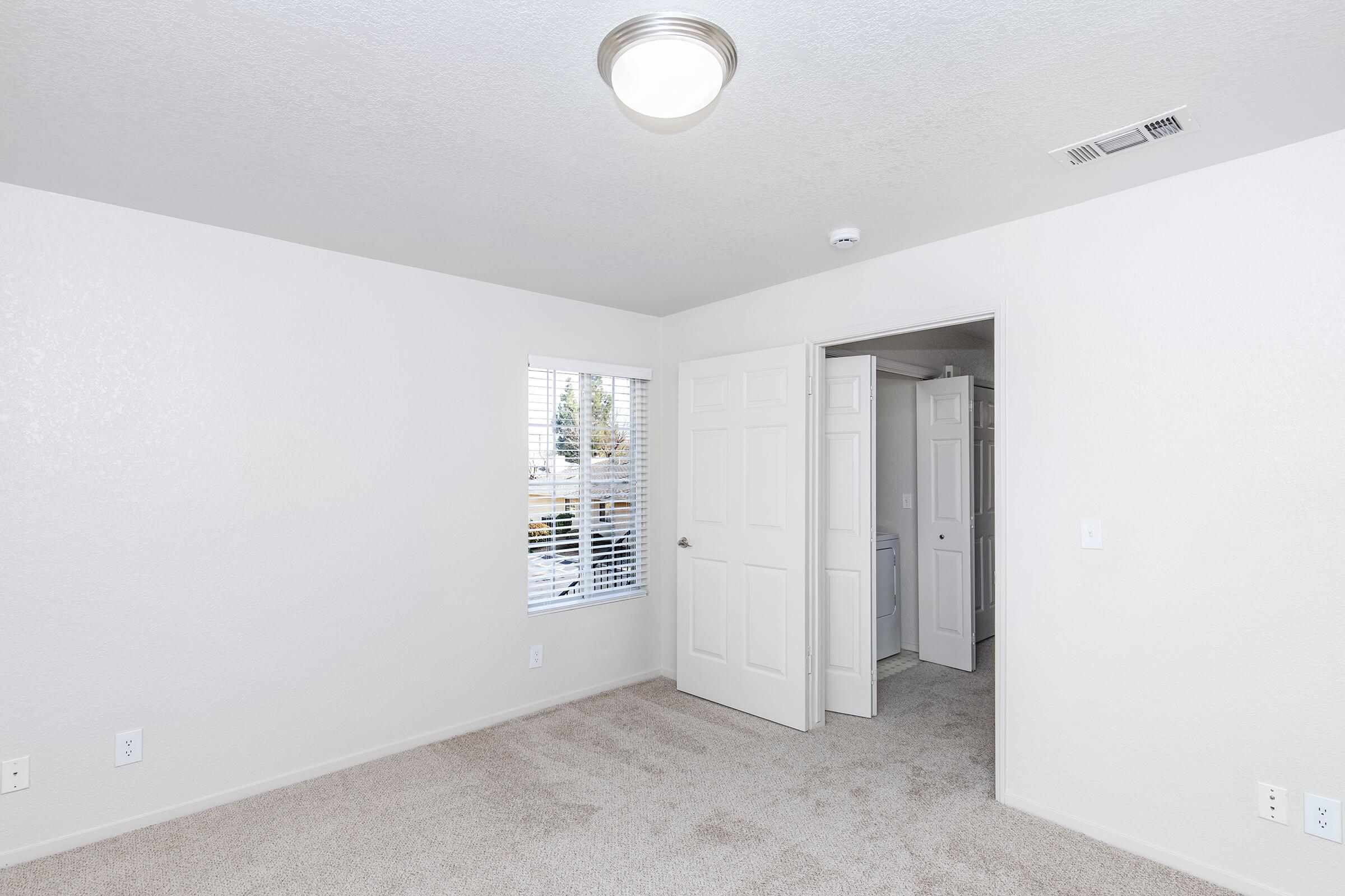 A bright, empty room with light gray carpet and white walls. It features a ceiling light fixture, a window with blinds, and an open door leading to another area. The space appears clean and ready for furniture, with additional doors visible in the background.