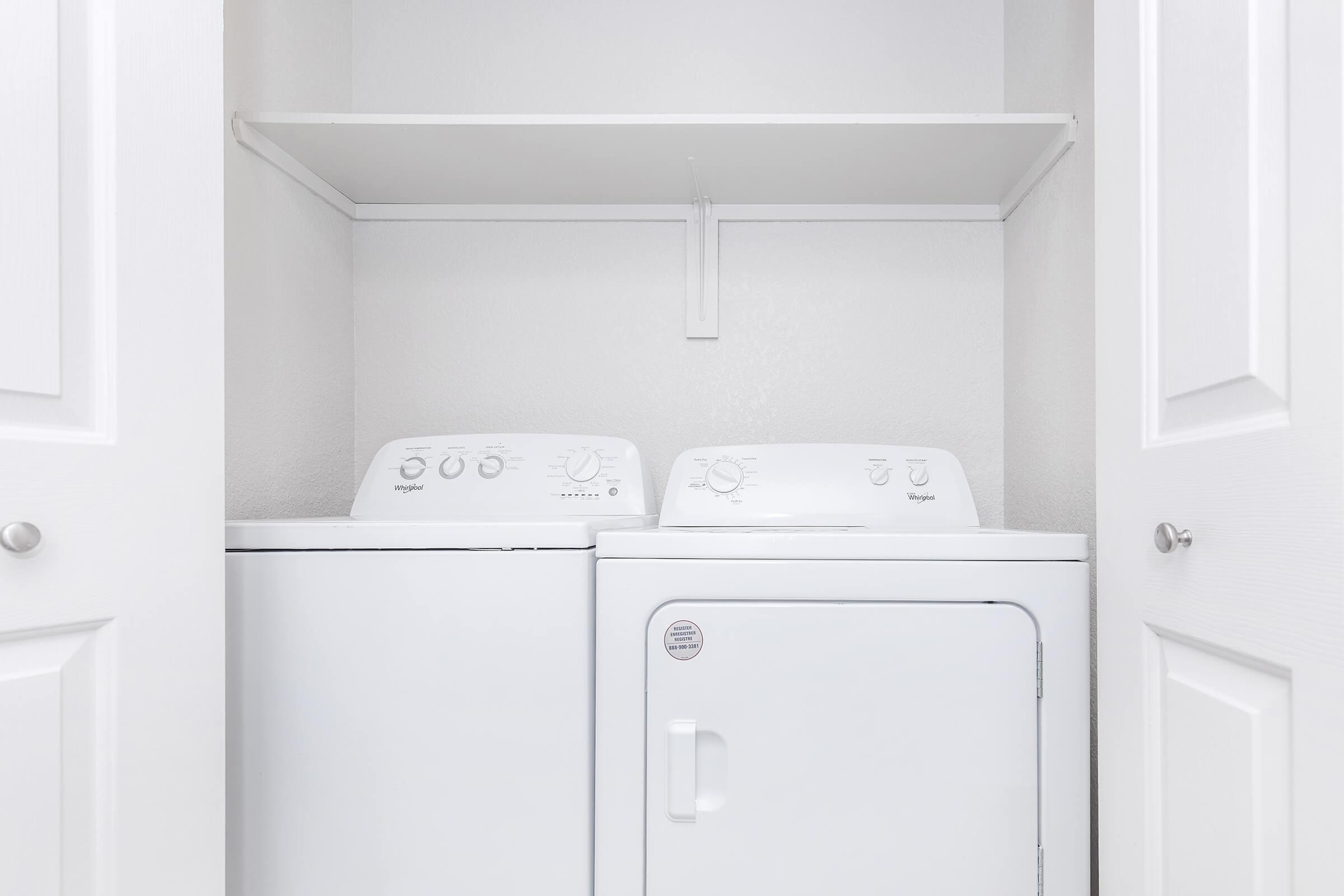 A compact laundry area featuring a white washing machine and dryer stacked side by side, with a shelf above. The walls are painted light gray, and the doors are closed, providing a clean and organized look. The appliances have simple dials and a modern design suitable for small spaces.