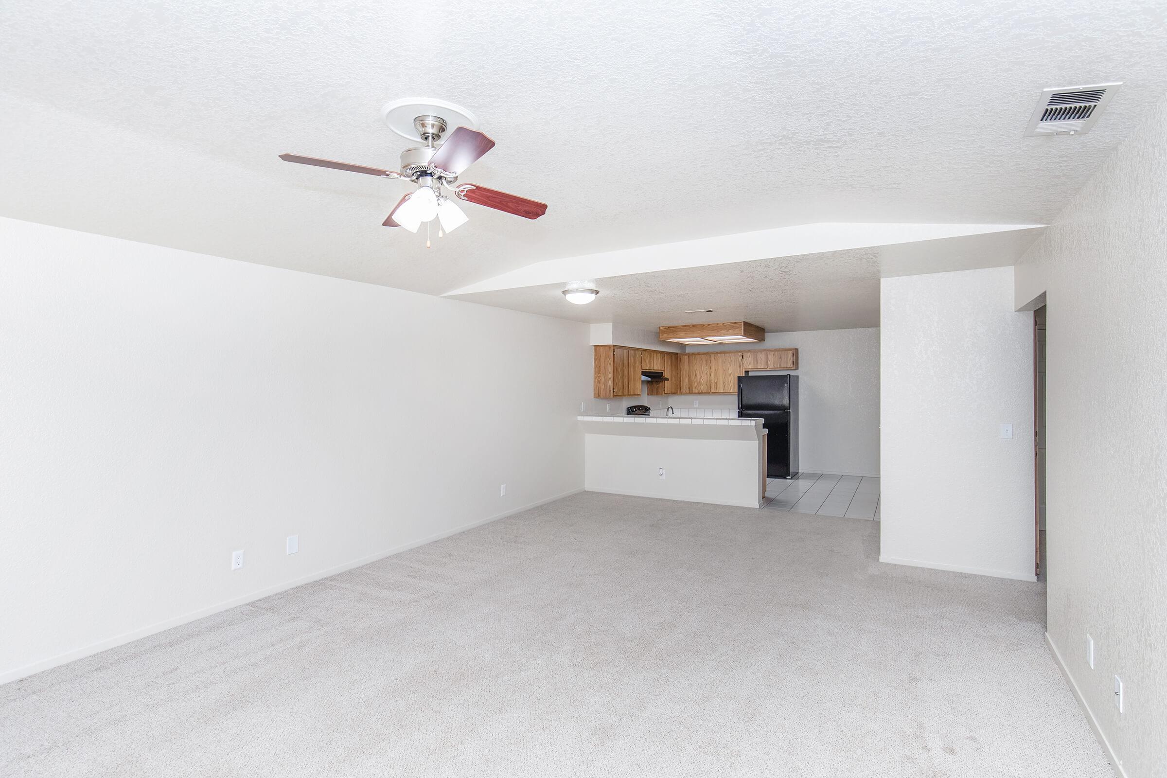 An empty living room with light-colored carpet and textured white walls. A ceiling fan is visible, and there is an open space leading to a kitchen area with wooden cabinets and a black refrigerator. The room is well-lit with natural light and has a simple, clean design.