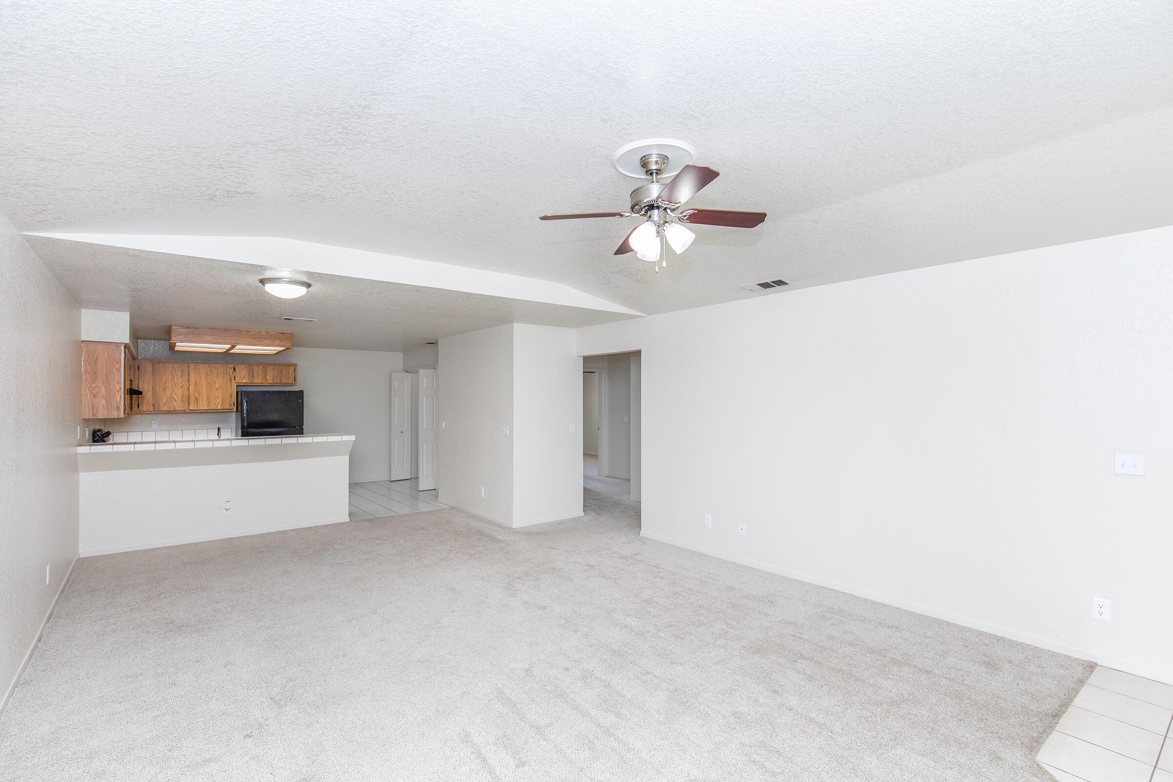 Spacious living area with light-colored carpet and a ceiling fan. The open floor plan connects to a kitchen area with wooden cabinets and a bar counter. A doorway leads to another room, while walls are painted in neutral tones, creating a bright and airy atmosphere.