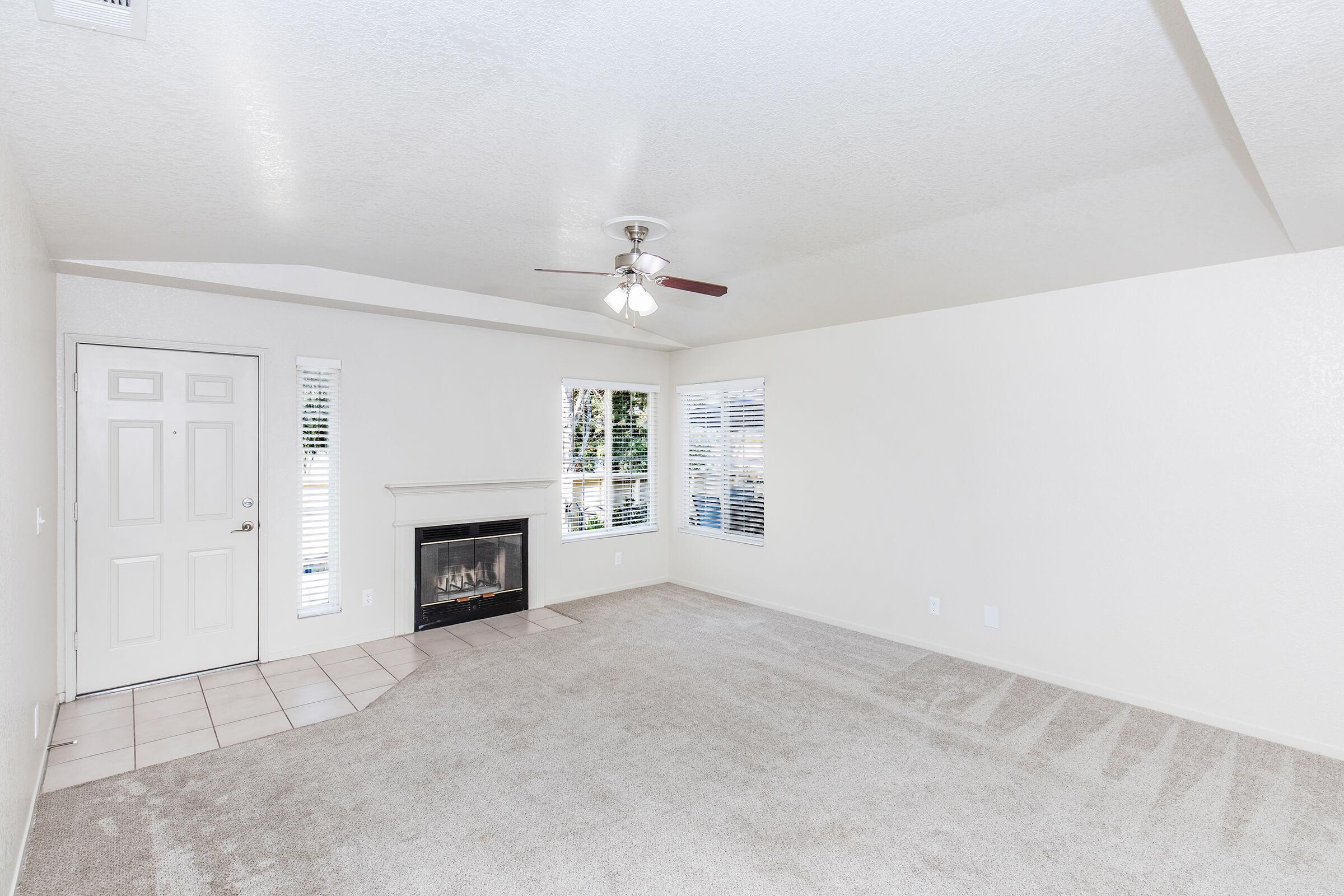 A spacious, empty living room featuring light beige walls, plush carpet, and a ceiling fan. There is a fireplace on the left side and a front door with a window beside it. Natural light filters in through two windows with blinds. The room has a clean and bright ambiance, ready for personalization.