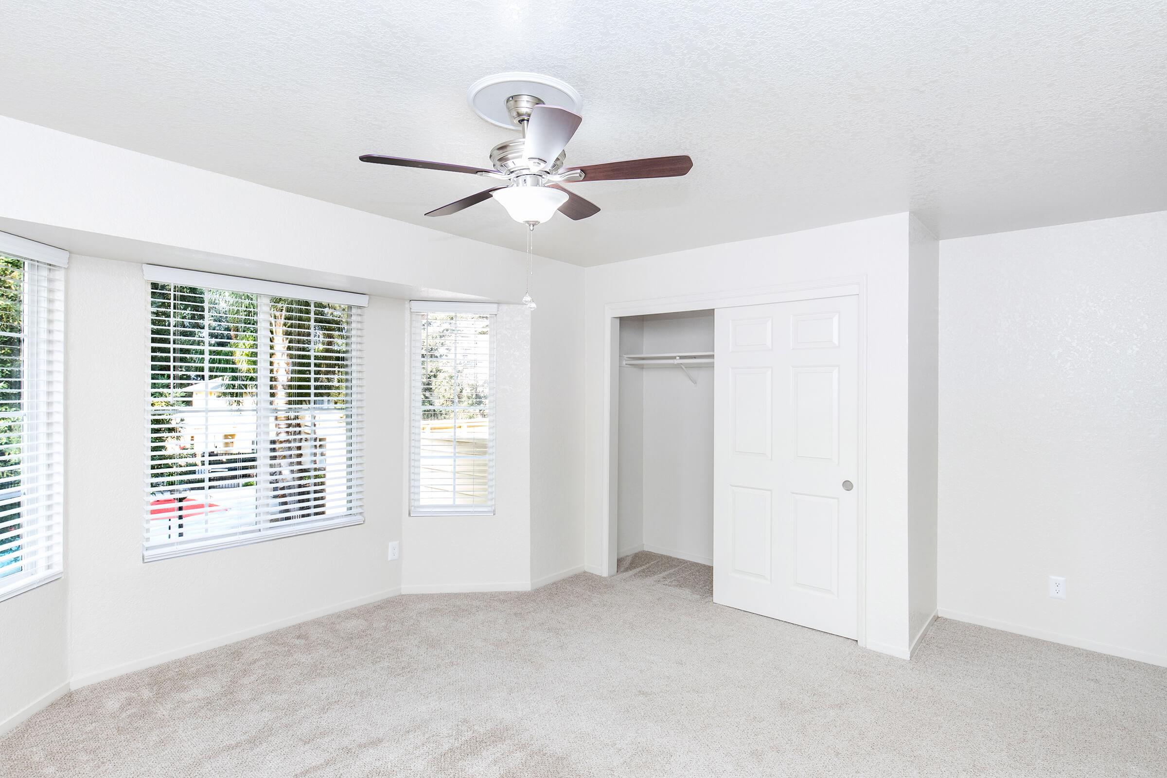 A bright, empty room with beige carpet, featuring a ceiling fan, large windows adorned with blinds, and a closet with sliding doors. The walls are painted white, creating a clean and spacious atmosphere. Natural light fills the space, enhancing its openness.