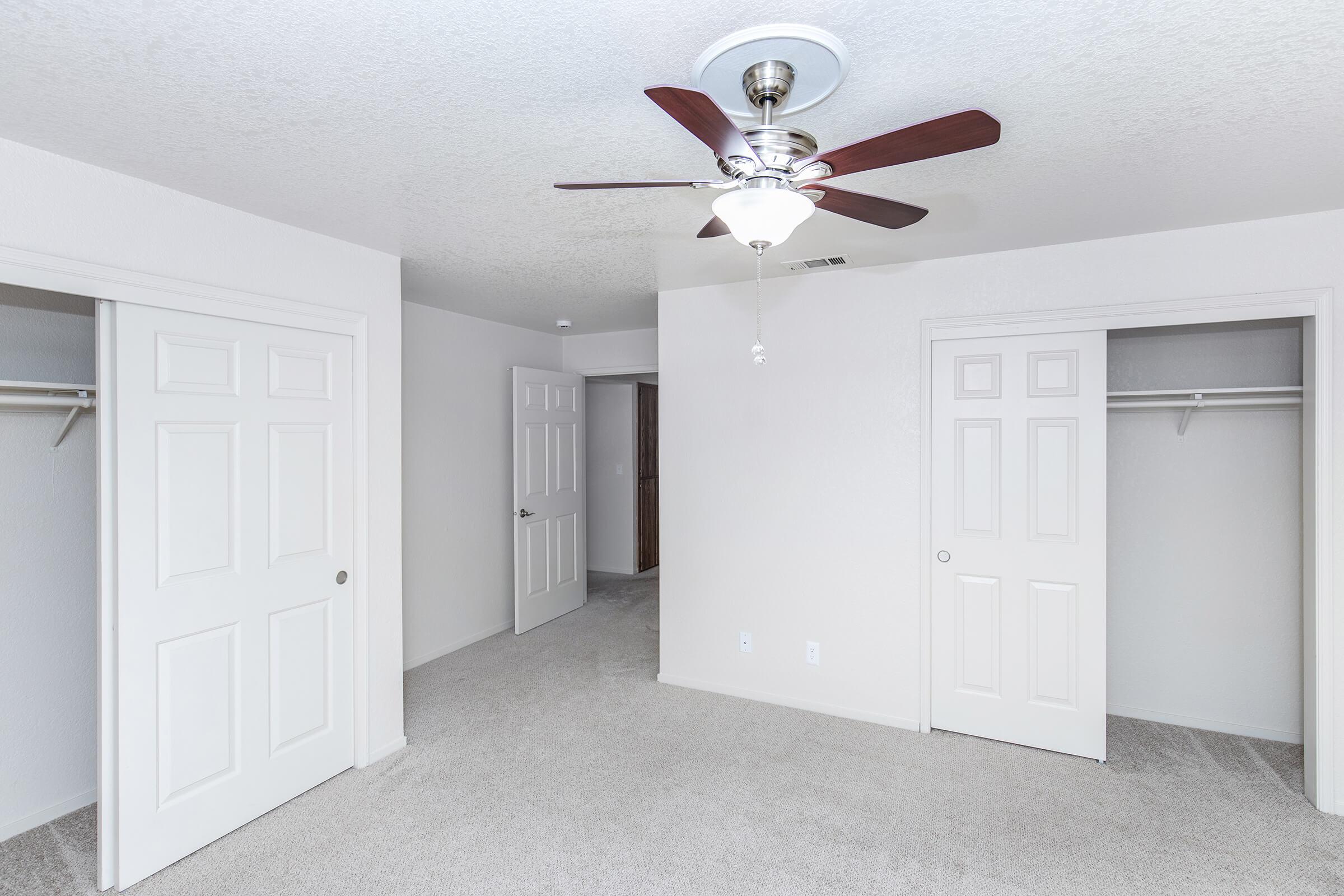 A spacious, empty room with light-colored walls and carpet. It features a ceiling fan with wooden blades, two closed doors leading to closets, and a doorway leading to another room. The overall ambiance is bright and airy, with natural light illuminating the space.