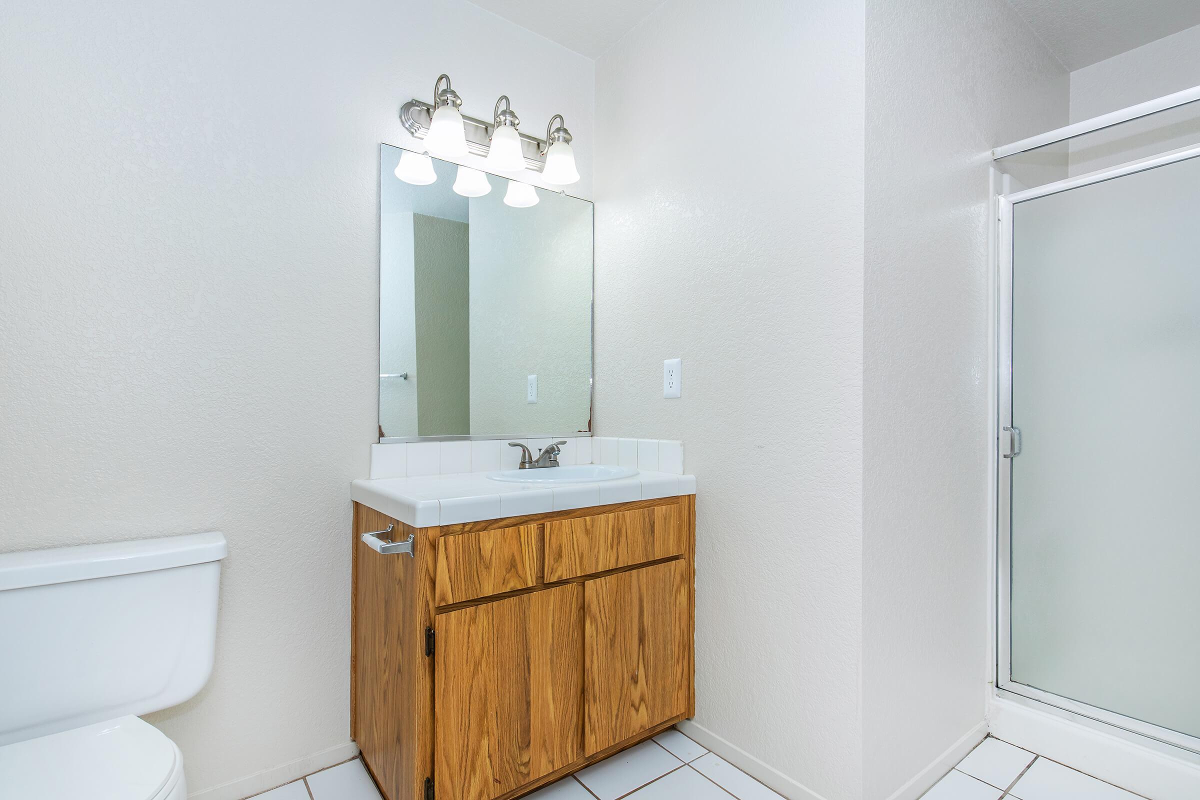 A clean, modern bathroom featuring a wooden vanity with a sink and two drawers, a large mirror above the vanity, a toilet, and a glass-enclosed shower. The walls are painted in a light color, and the floor is tiled with white tiles. Bright lighting is provided by three wall-mounted light fixtures.