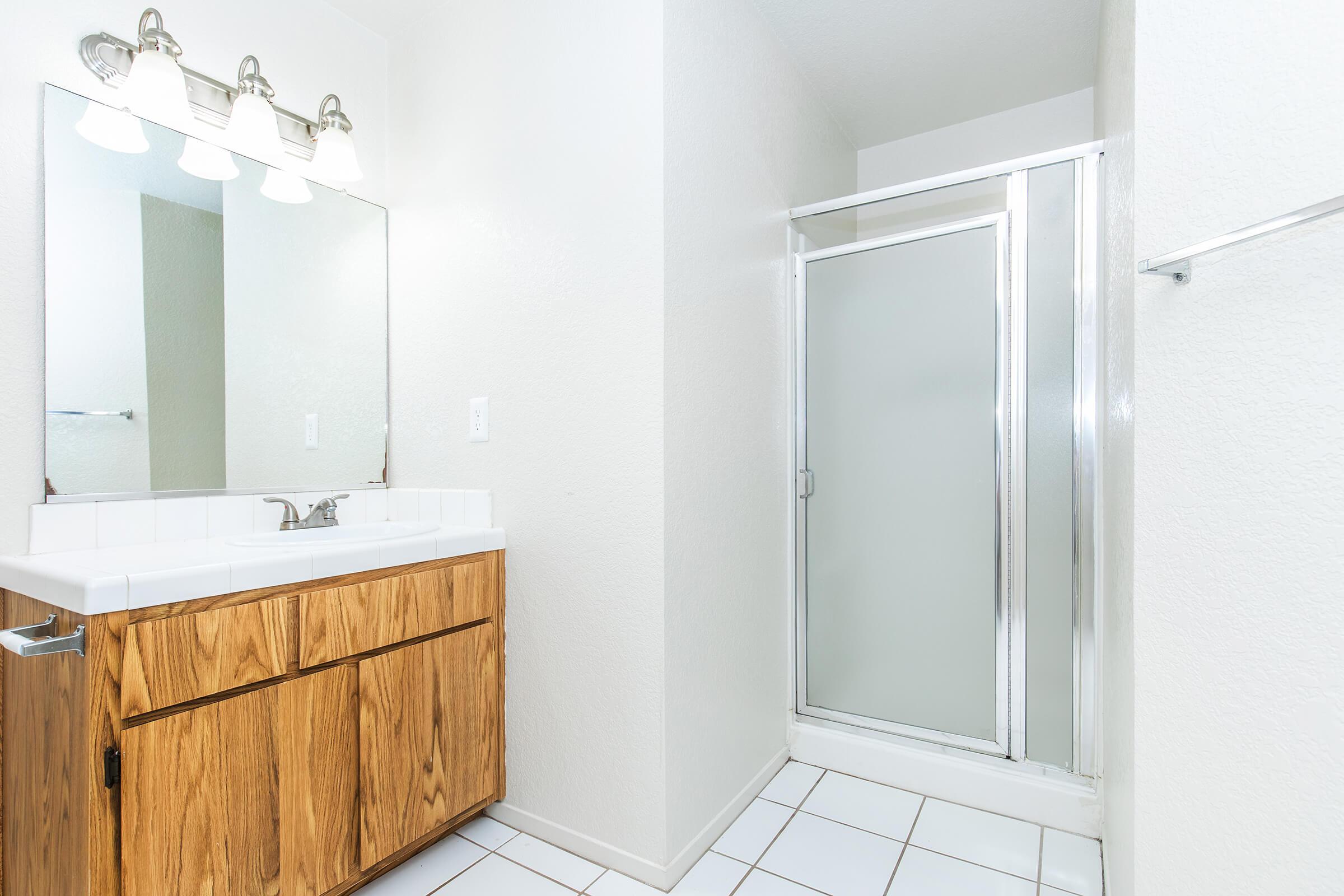 A modern bathroom featuring a large mirror above a white countertop with wooden cabinets below. A sliding glass shower door is visible in a separate corner. The walls are painted white, and the floor is tiled with white square tiles. Bright lighting illuminates the space.