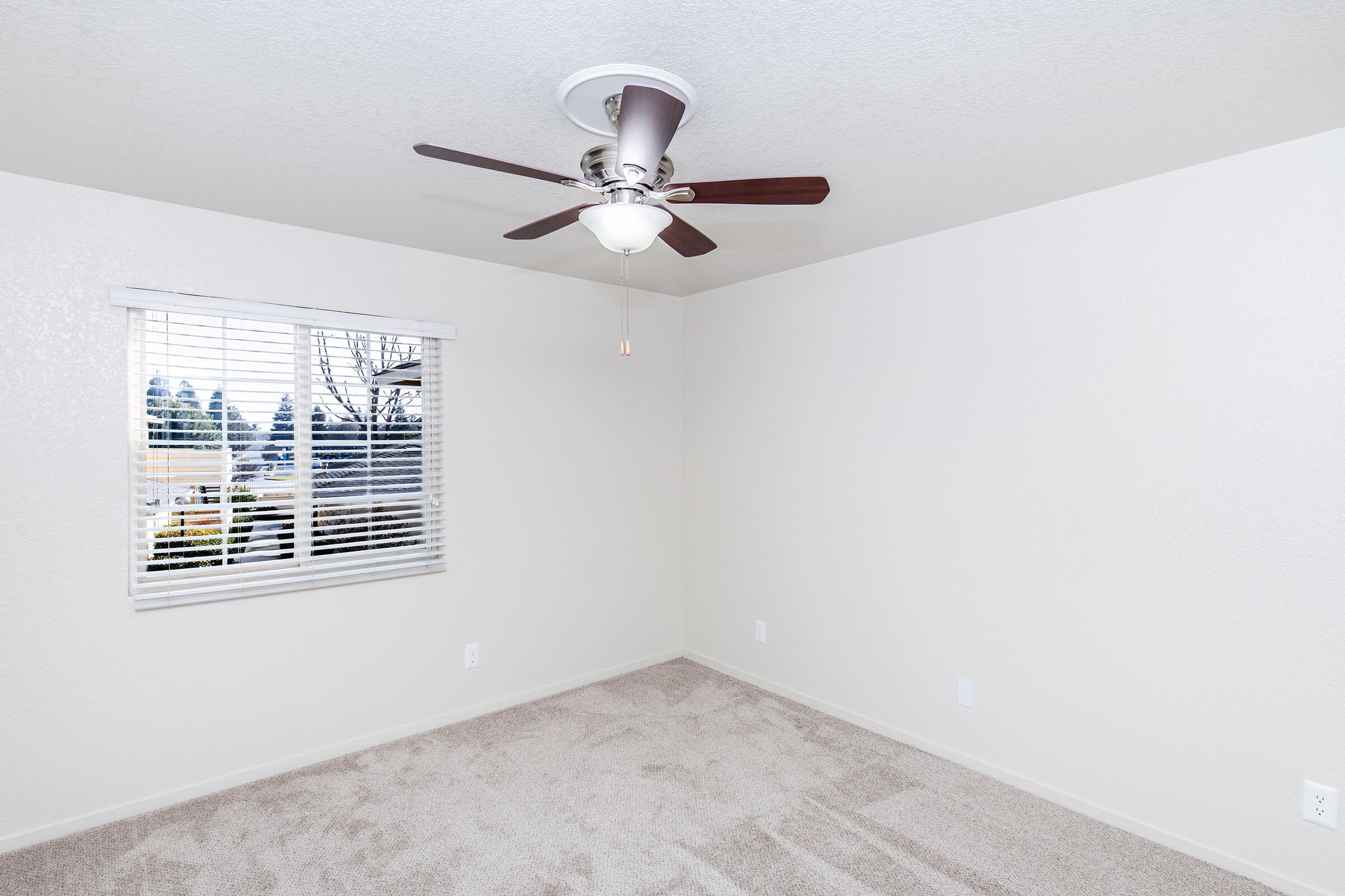 A bright, empty room with light beige walls and a ceiling fan with wooden blades. There’s a window with blinds on one wall, allowing natural light to enter. The floor is covered in soft, light-colored carpet, creating a warm and inviting atmosphere.