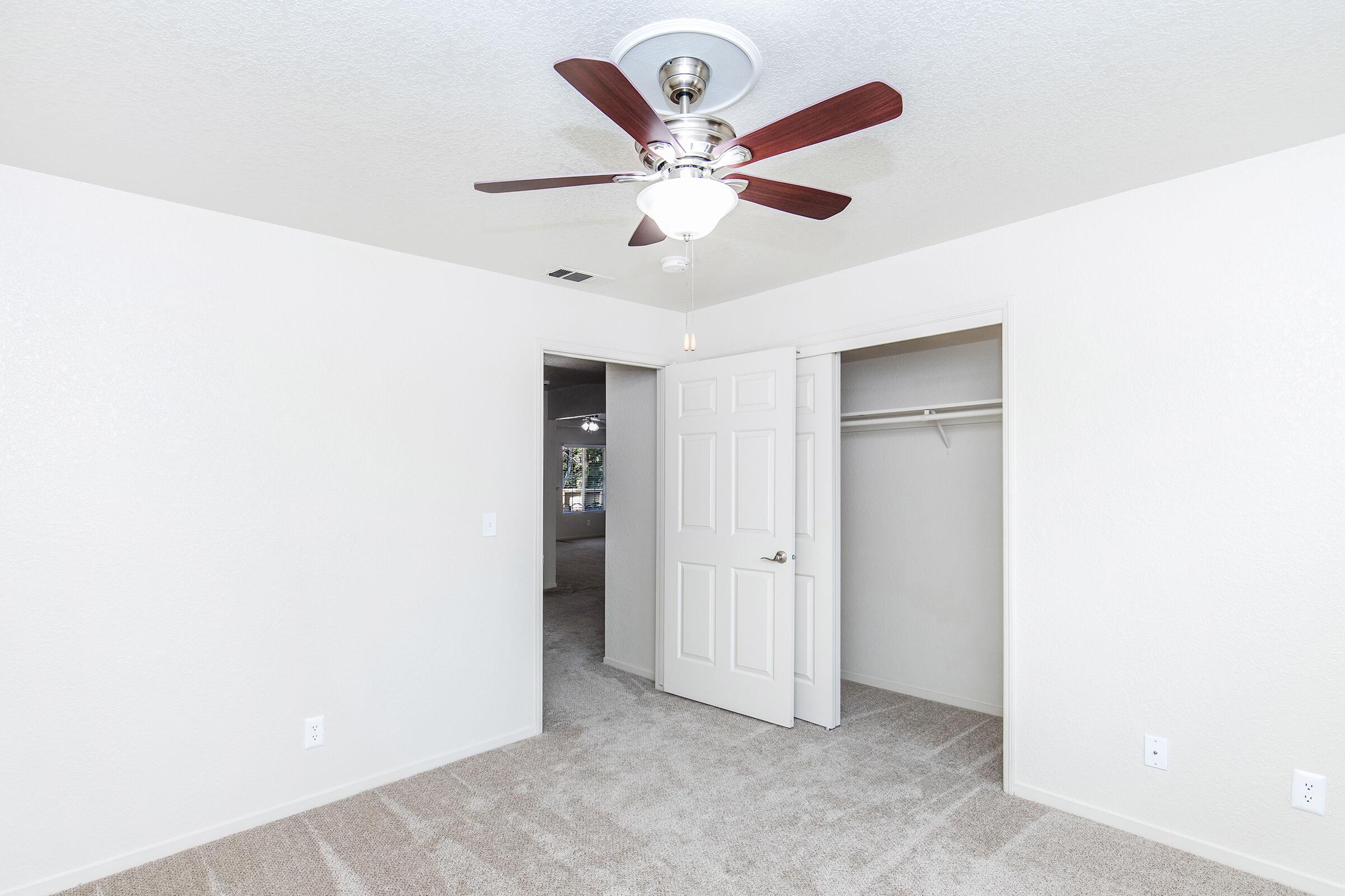 A well-lit bedroom featuring beige walls, a ceiling fan with wooden blades, and plush carpet. The room has an open door leading to a closet space and another room in the background. Natural light streams in, creating a warm and inviting atmosphere.