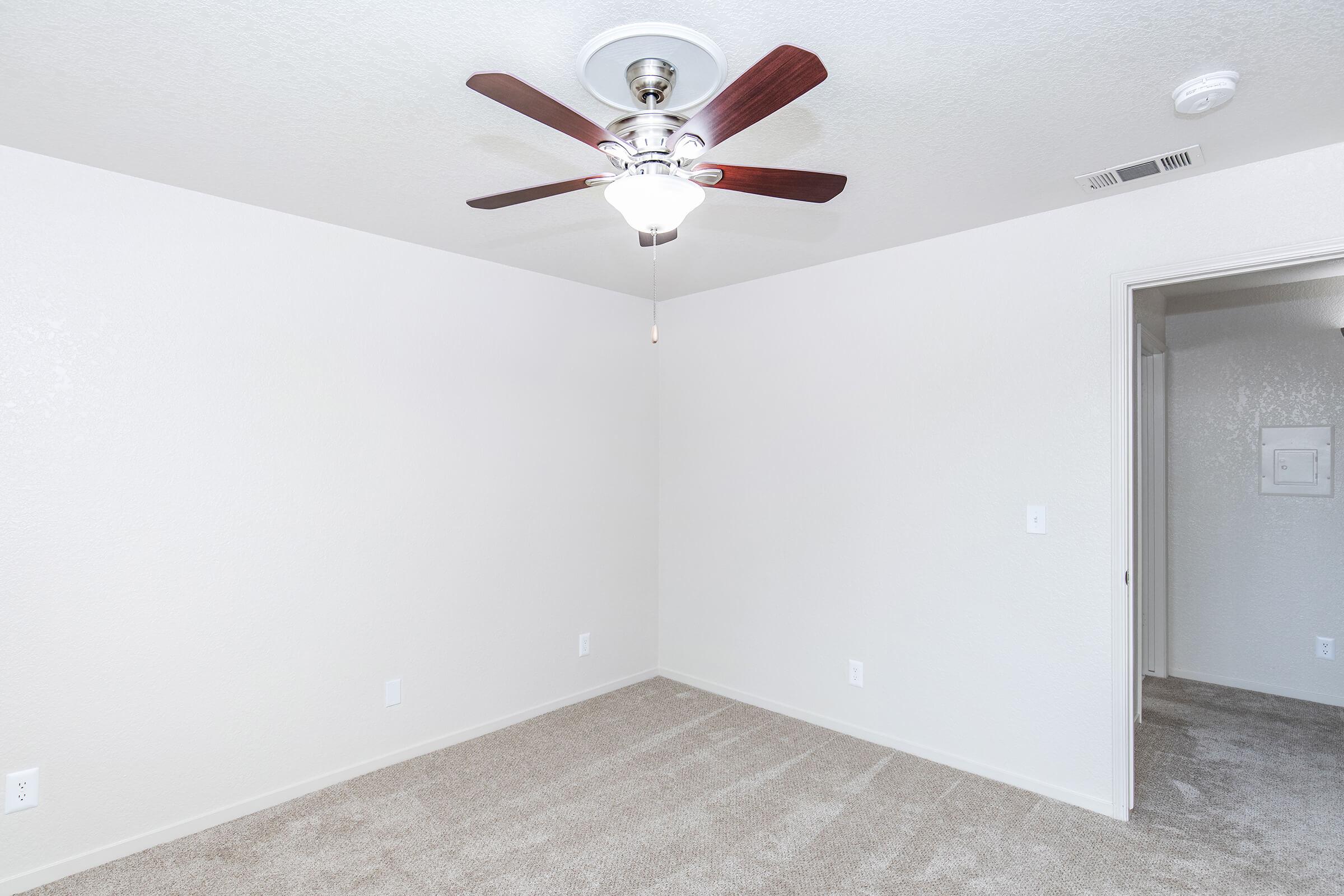 Empty room with light-colored walls and a ceiling fan featuring wooden blades. The floor is covered in beige carpet, and there is a doorway on the right leading to another space. The overall ambiance is bright and neutral, with no furniture present.