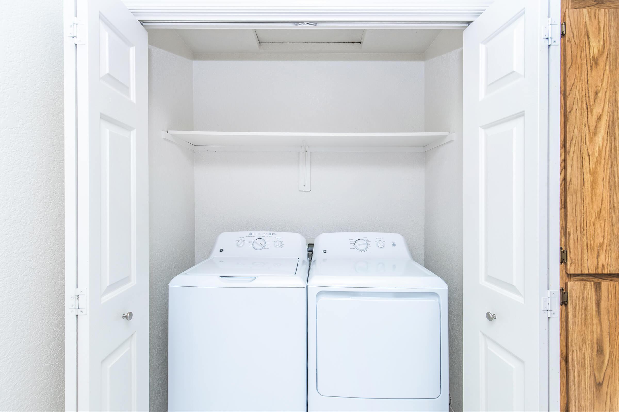 A stacked laundry setup featuring a washing machine and a dryer side by side, placed in a small closet space. The walls are light-colored, with a single shelf above the machines for storage. There is also wood cabinetry visible on the right side of the image.