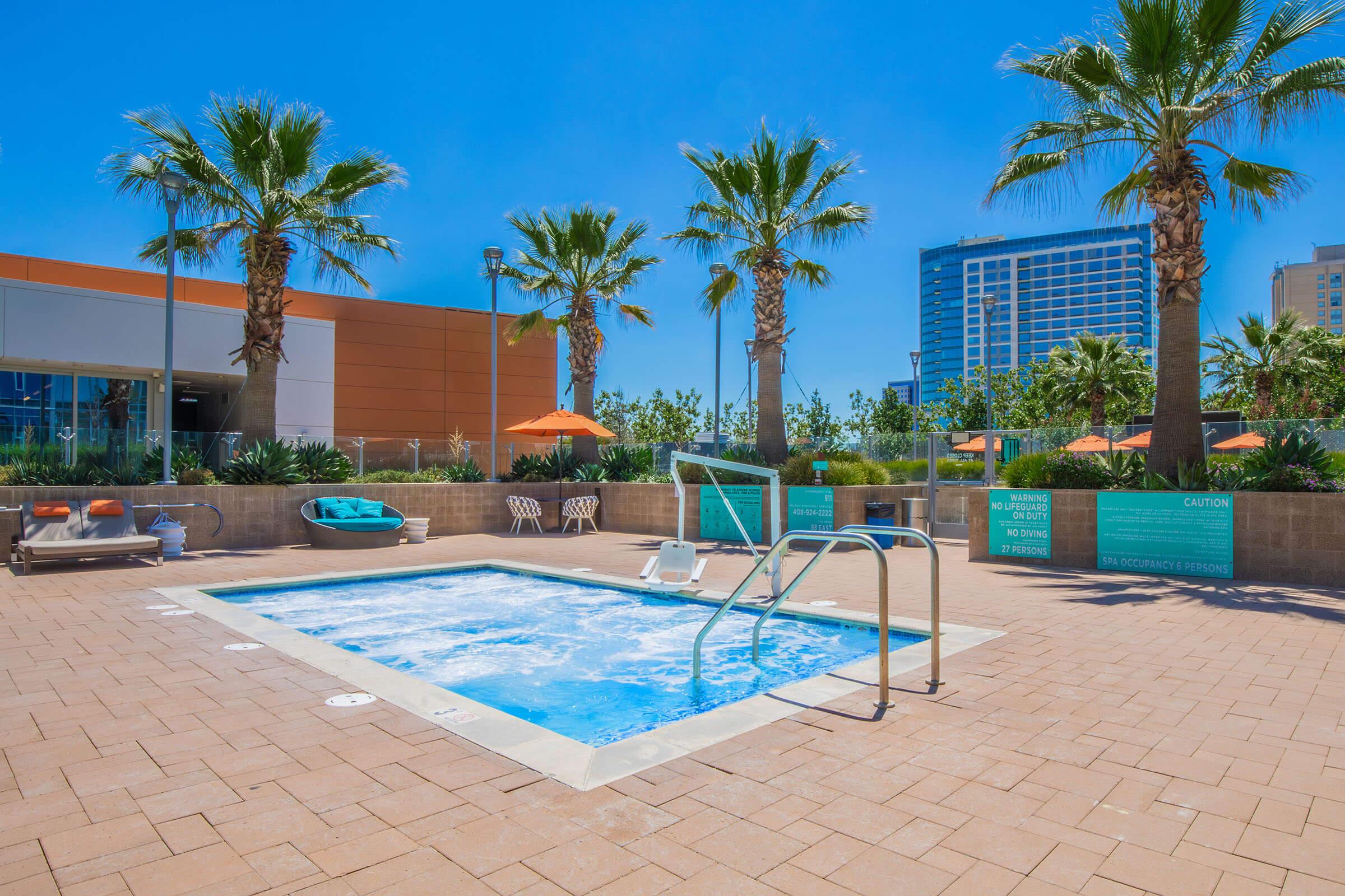 A clean, sunny pool area featuring a small hot tub surrounded by palm trees. There are lounge chairs and umbrellas for relaxation, as well as a light blue poolside area. The background includes a modern building, creating a vibrant and inviting atmosphere.