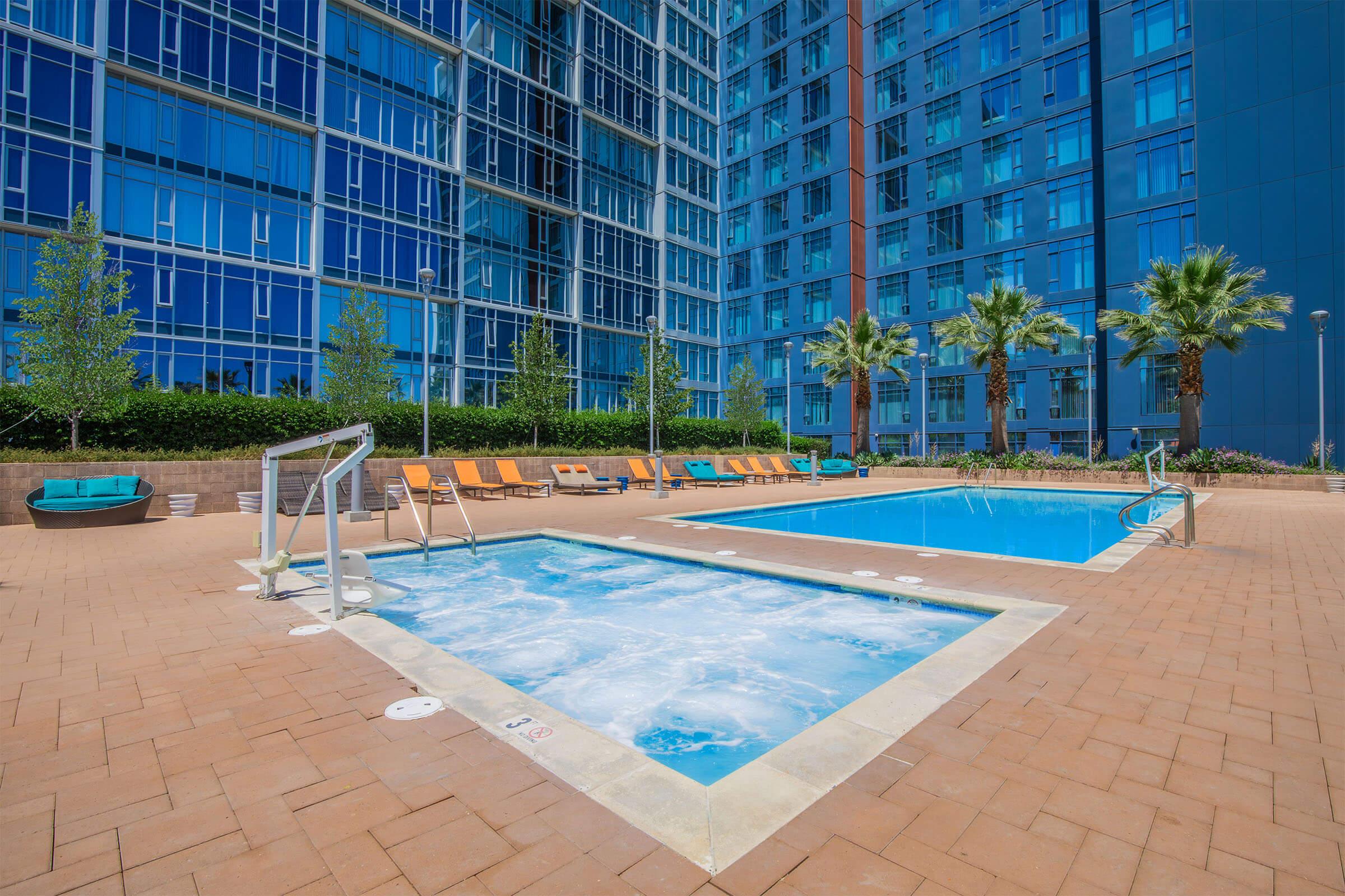 A bright outdoor swimming pool area featuring a large pool and a smaller hot tub. Surrounding the pools are lounge chairs in vibrant colors, with palm trees and landscaped greenery in the background. The modern building's reflective glass facade adds a contemporary touch to the setting.