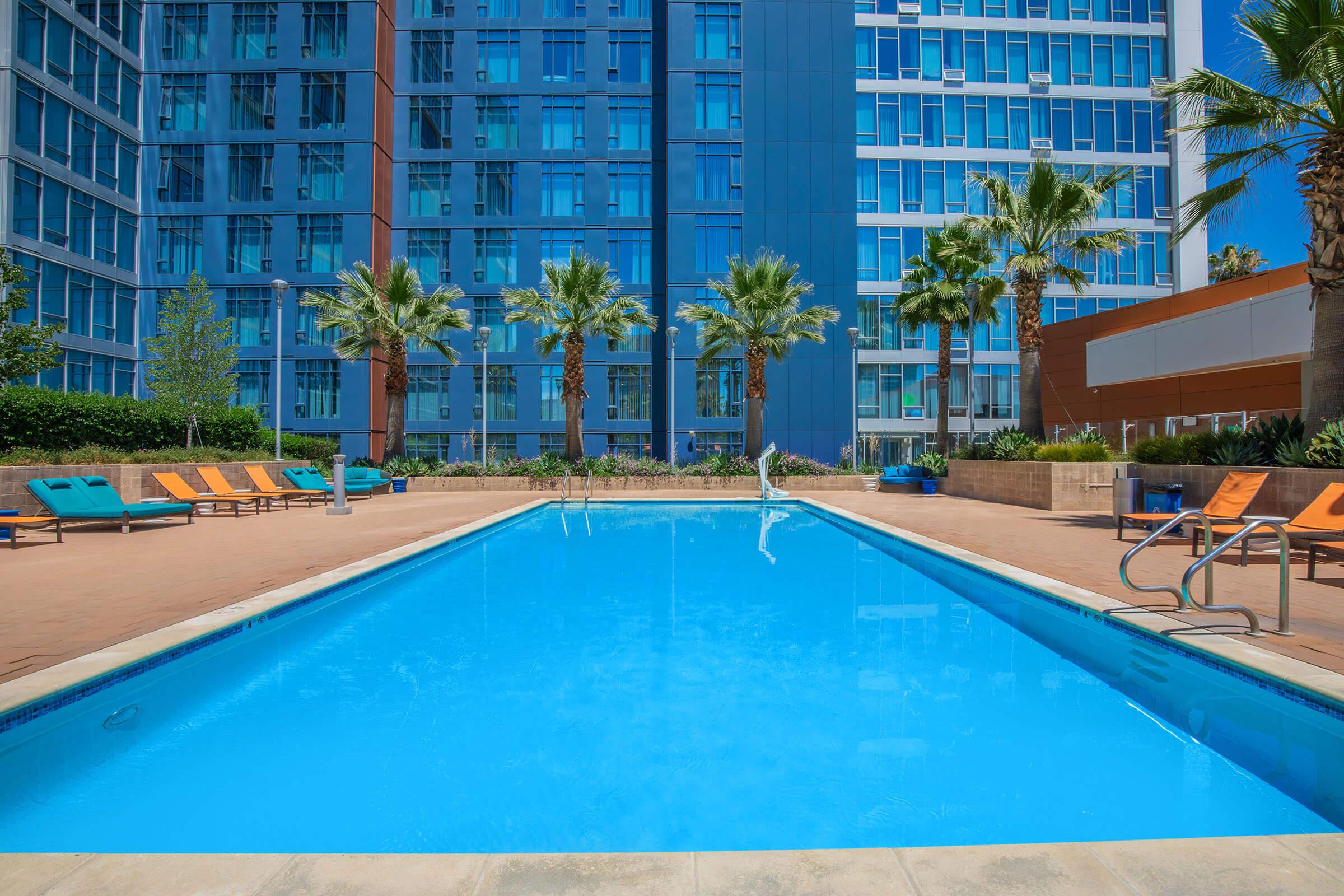 Outdoor swimming pool surrounded by comfortable lounge chairs and palm trees, with modern apartment buildings in the background. The pool features clear blue water and is set in a well-maintained landscape.