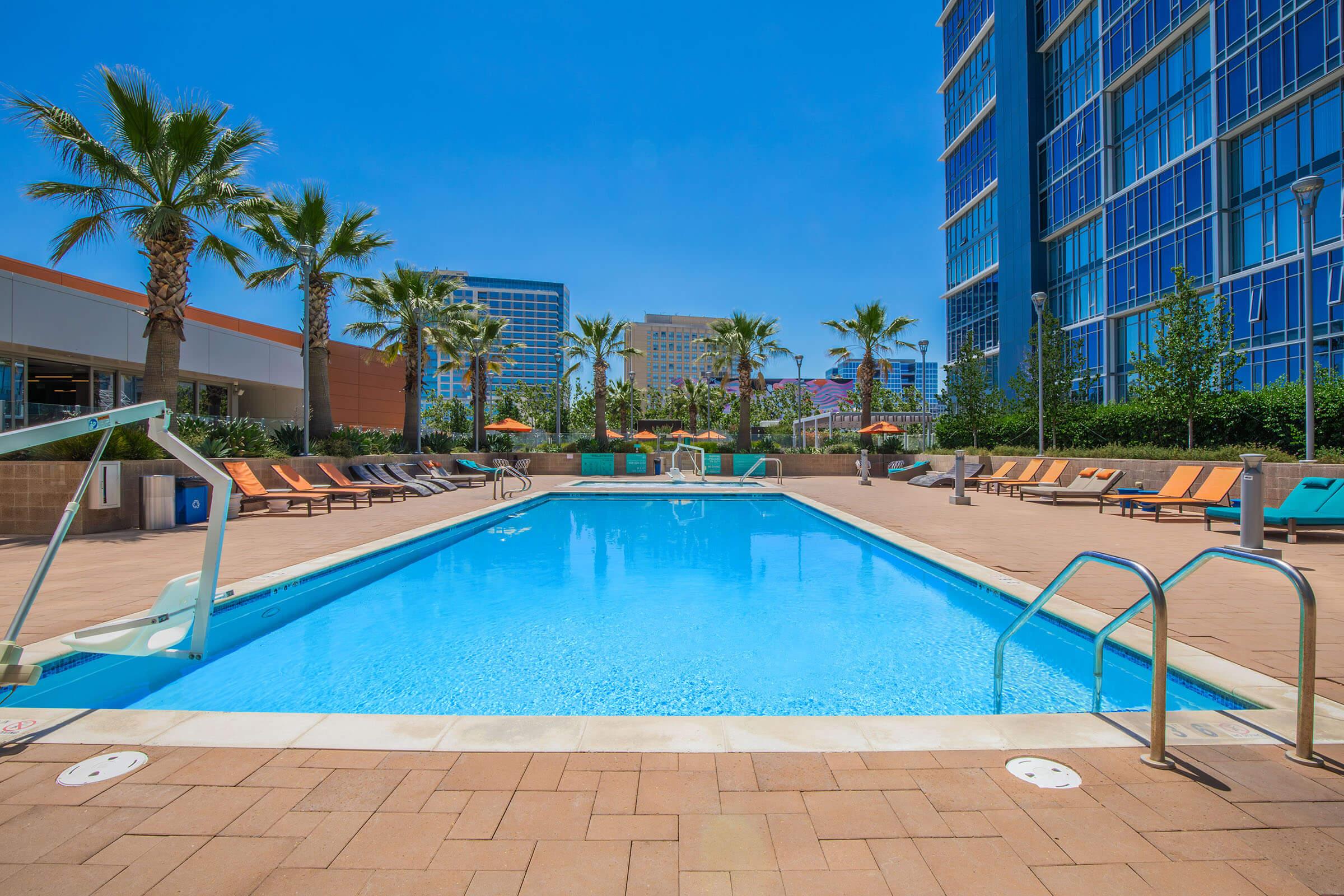A spacious outdoor pool surrounded by palm trees and lounge chairs. The clear blue water reflects the bright sky, with modern buildings in the background. Umbrella-shaded tables provide seating, creating a relaxing atmosphere in a sunny environment.