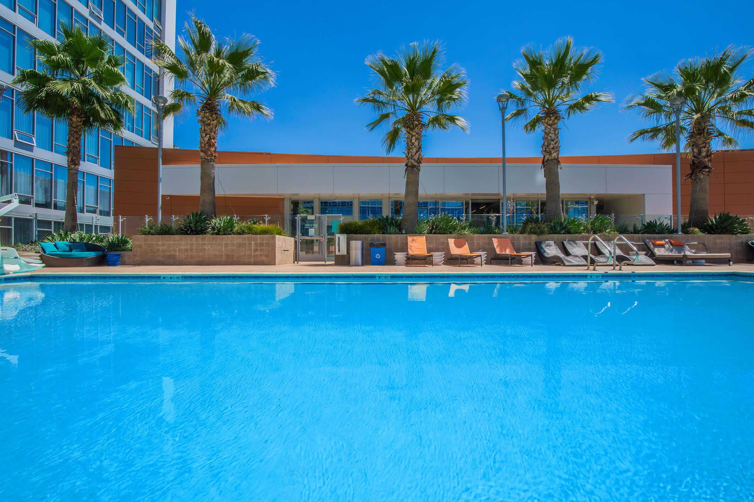 A sparkling blue swimming pool in front of a modern building, surrounded by lounge chairs and palm trees. The clear sky and bright sunshine enhance the vibrant atmosphere, creating a relaxing outdoor space for leisure and enjoyment.