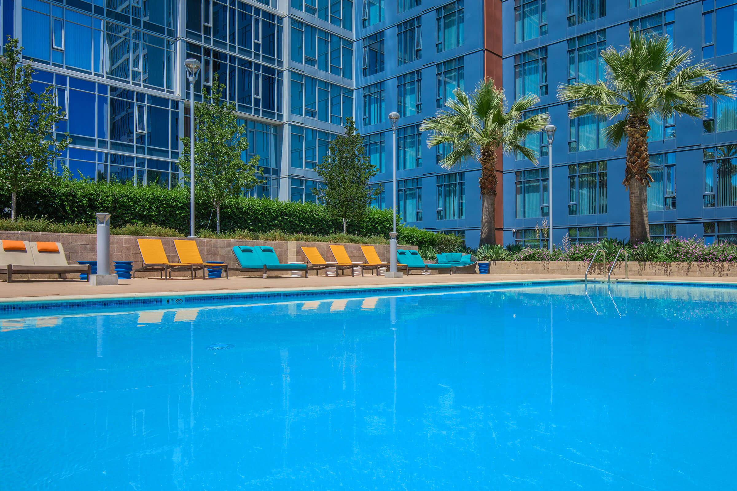 A clear blue swimming pool surrounded by lounge chairs in vibrant colors, with tall palm trees and modern glass buildings in the background. The scene captures a sunny, relaxing atmosphere, ideal for leisure and enjoyment.