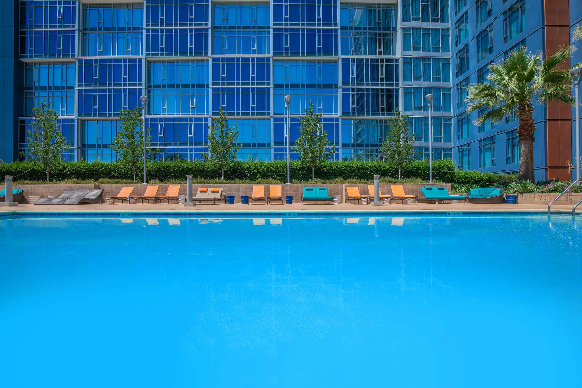 A clear blue swimming pool in the foreground, with lounge chairs arranged along the edge. In the background, a modern building with large blue-tinted windows and neatly trimmed greenery. Palm trees are visible near the pool area, creating a relaxed outdoor atmosphere.