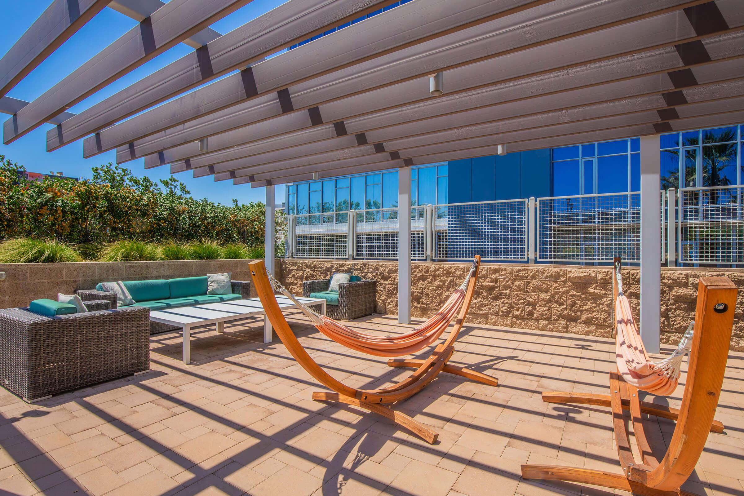 A peaceful outdoor space featuring two wooden hammocks, a modern seating area with cushions, and a large table under a slatted pergola. The background shows green foliage and a blue-glass building, creating a relaxing atmosphere for lounging or socializing.