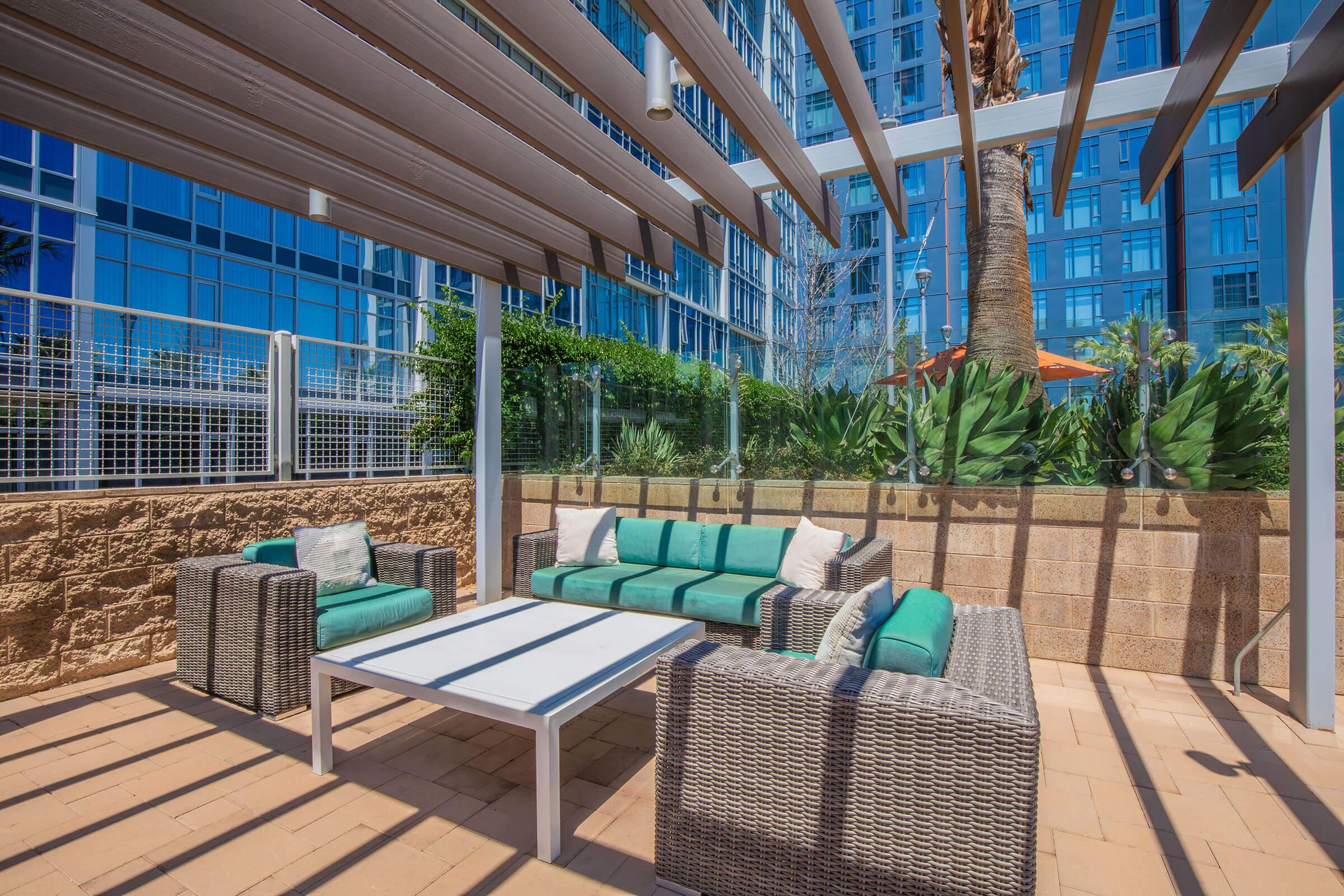 A modern outdoor lounge area featuring a white table and comfortable seating with teal cushions. Surrounded by greenery and sleek glass buildings, the space is shaded by a pergola, creating a relaxing environment ideal for socializing or enjoying the outdoors.