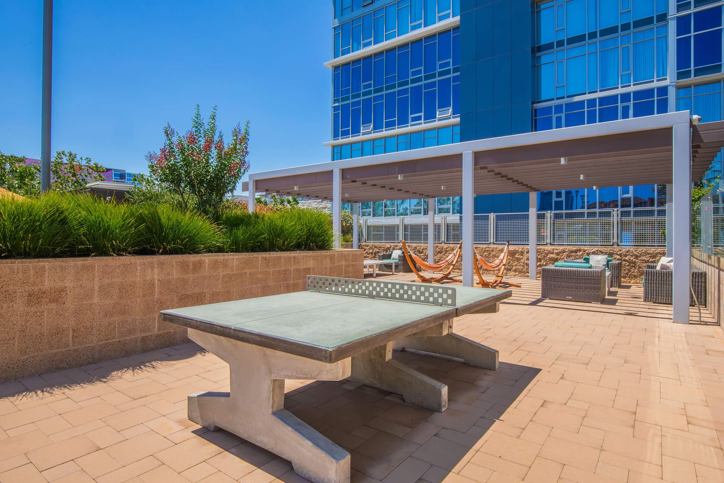 Outdoor recreational area featuring a concrete ping pong table, surrounded by lush greenery and modern seating options. In the background, a sleek glass building is visible under a clear blue sky, alongside shaded lounging spaces with hammocks.