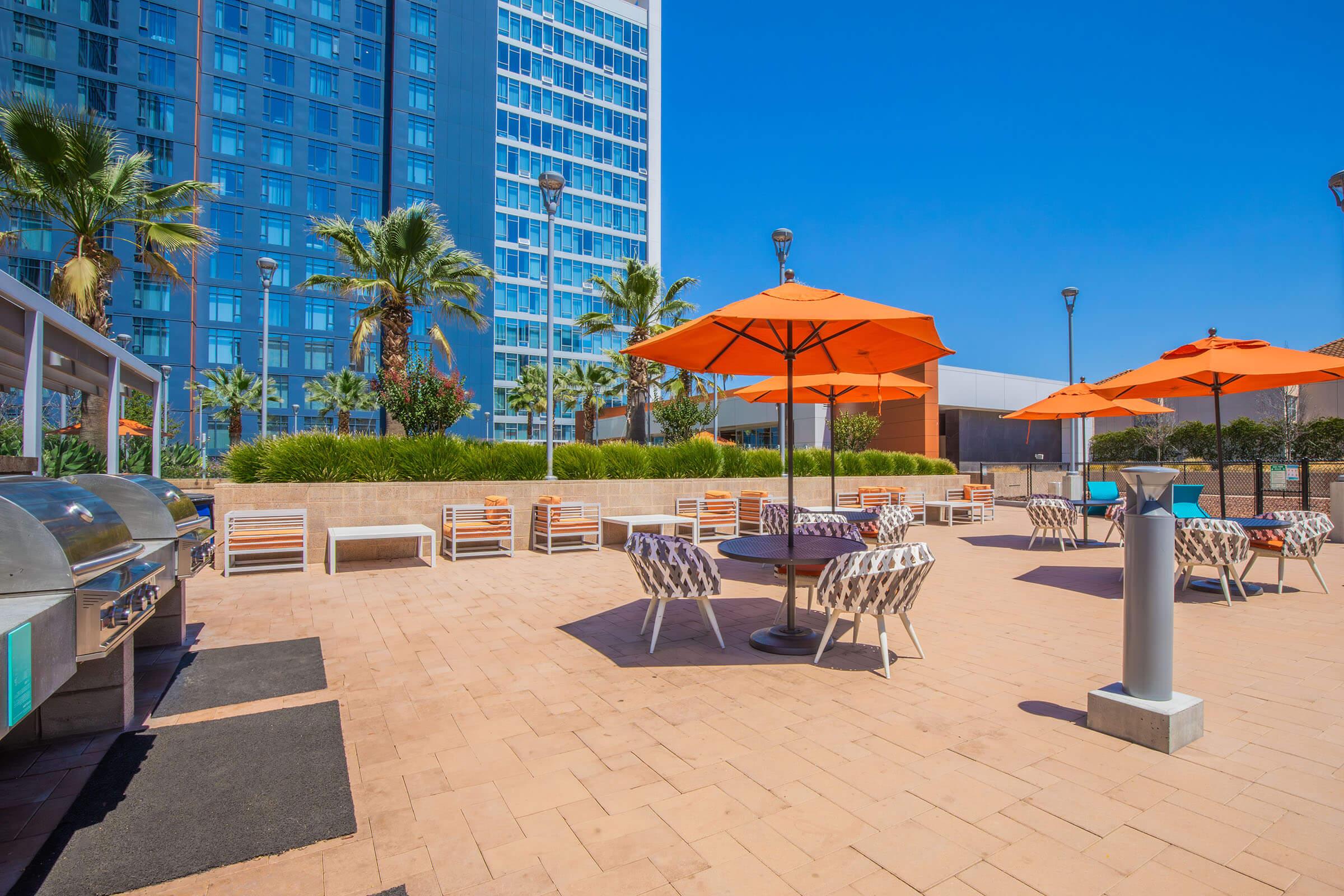 An outdoor patio area features several orange umbrellas providing shade over tables and chairs. There are grilling stations and palm trees in the background, with a modern high-rise building visible nearby under a clear blue sky. The space is designed for relaxation and social gatherings.
