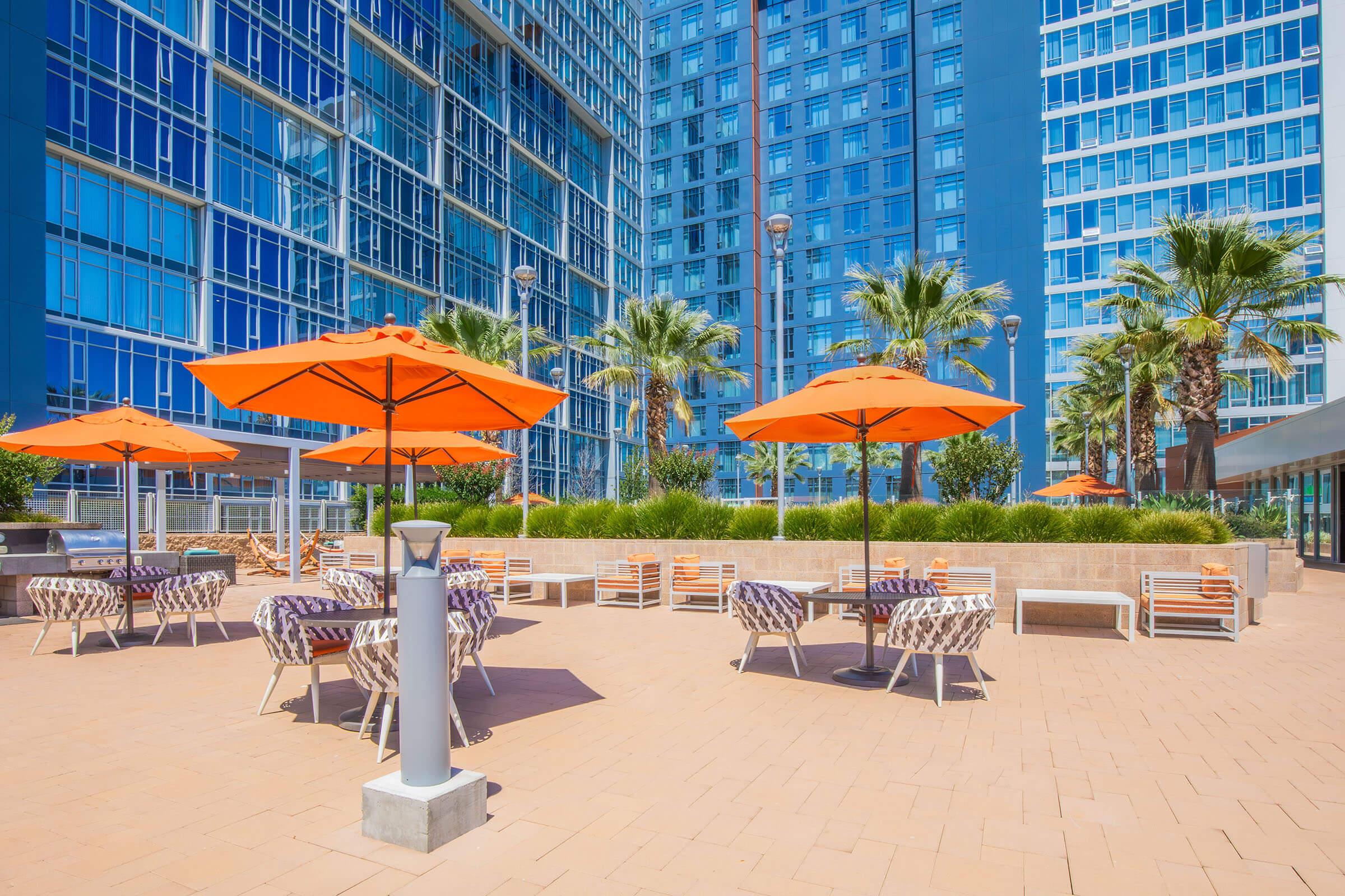 A modern outdoor patio area featuring several lounge chairs and tables with orange umbrellas. Surrounding the space are tall buildings with large glass windows and tropical palm trees, creating a vibrant and inviting atmosphere. Sunlight fills the area, enhancing the lively ambiance.