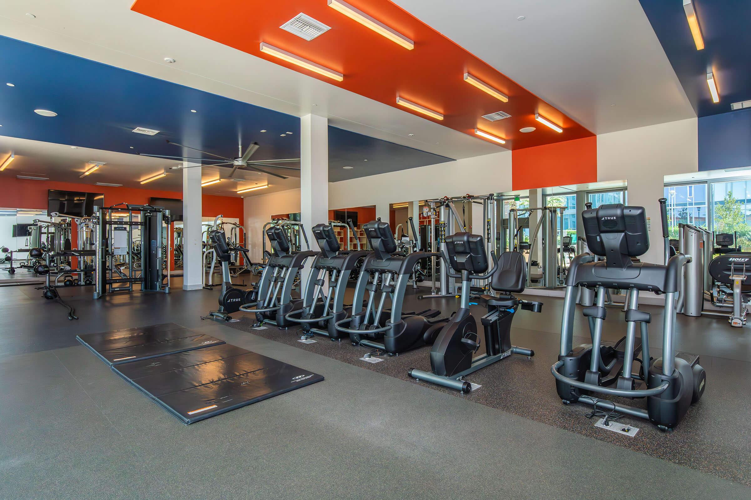 A modern gym interior featuring various exercise machines, including elliptical trainers and weightlifting equipment. The space is well-lit with colorful walls, mirrors, and a clean, organized layout. Fitness mats are positioned on the floor, creating an inviting atmosphere for workouts.