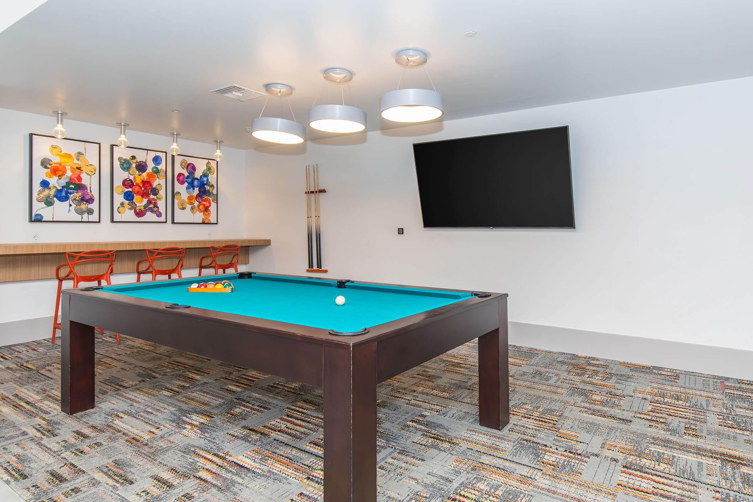 A game room featuring a pool table with a teal felt surface, illuminated by modern light fixtures. The wall is decorated with colorful artwork. Behind the pool table, there are three red chairs and a large flat-screen TV mounted on the wall. The floor is covered with patterned carpet.