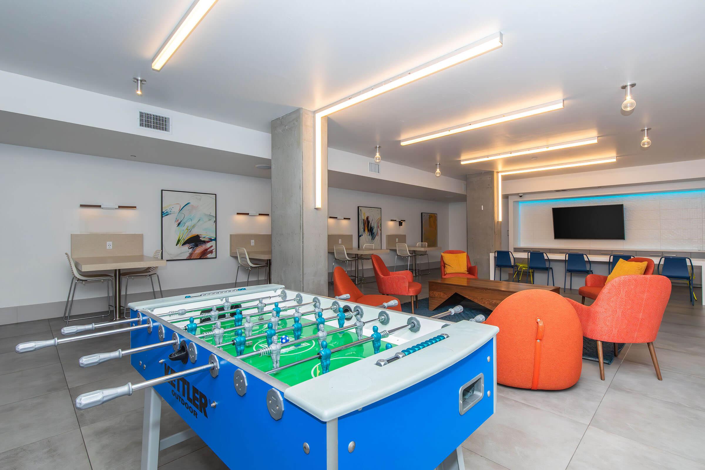 A modern lounge area featuring a foosball table in the foreground. The space includes colorful chairs, a coffee table, and wall art. Bright, overhead lighting illuminates the room, which has neutral-toned walls and flooring, creating a welcoming atmosphere for social gatherings.