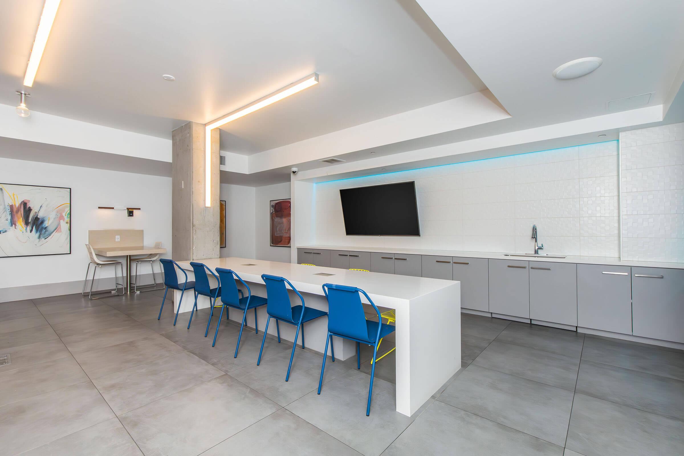 Modern kitchen space featuring a long white table with blue chairs. The room has sleek gray cabinetry and a large wall-mounted television. Soft lighting enhances the contemporary design, while a small kitchenette area with a sink is visible in the background.