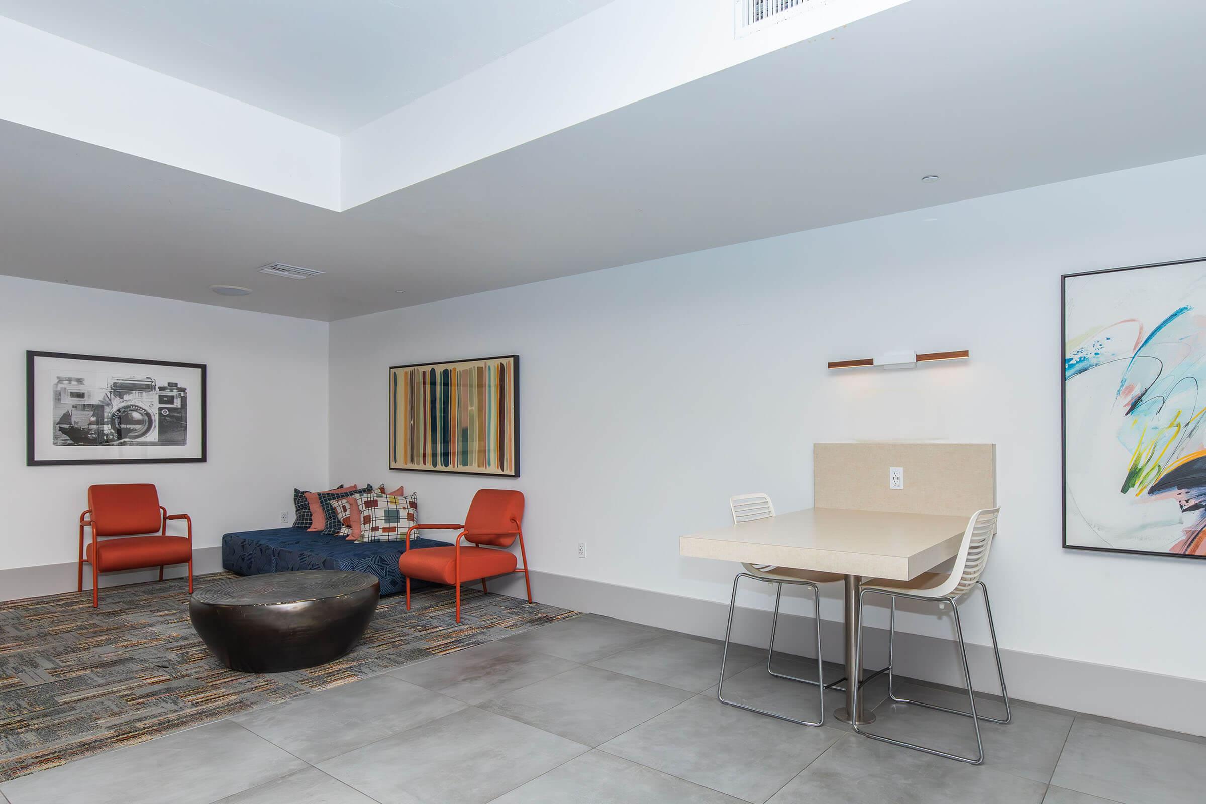 Modern lounge area with a seating arrangement featuring two orange chairs and a dark blue upholstered sofa with patterned cushions. A minimalist table and chairs are against a white wall adorned with abstract art. The floor is tiled, and a round, dark coffee table complements the space.