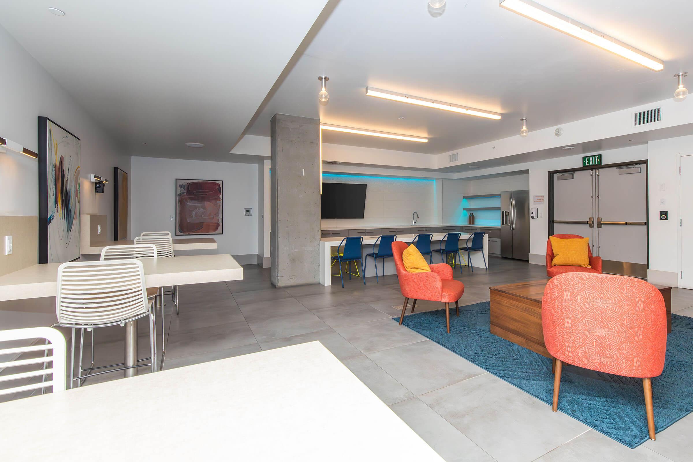 Modern communal space with a light, open atmosphere. Features a kitchen area with a large screen, tables with striped chairs, colorful accent chairs, and a wooden coffee table on a blue rug. Soft lighting from overhead fixtures enhances the inviting decor.