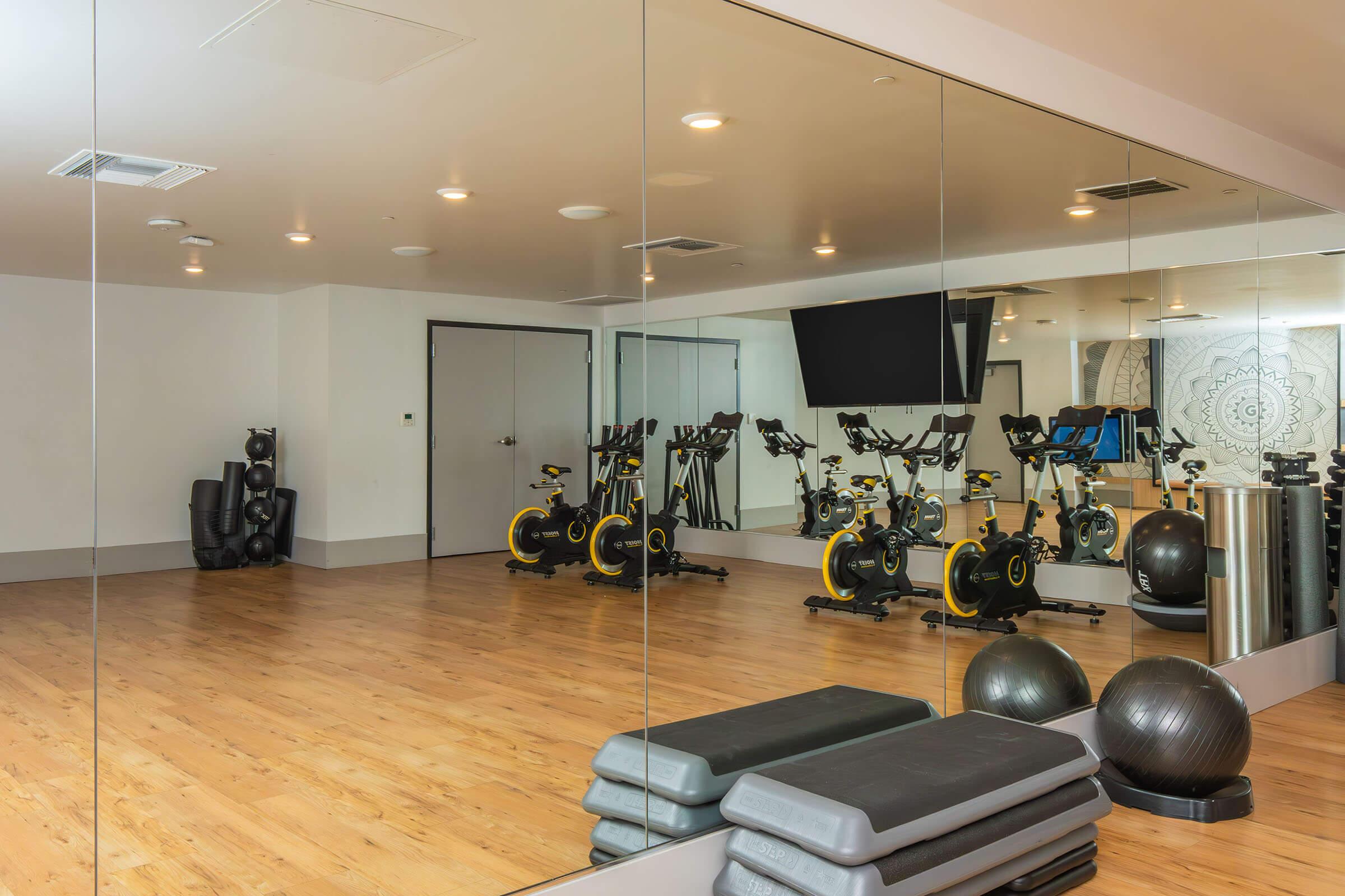 Interior of a fitness studio featuring several stationary exercise bikes arranged in front of a large mirror. The floor is wooden, and there are exercise mats and stability balls in the corner. A large screen is mounted on the wall, and the overall space is bright and modern.